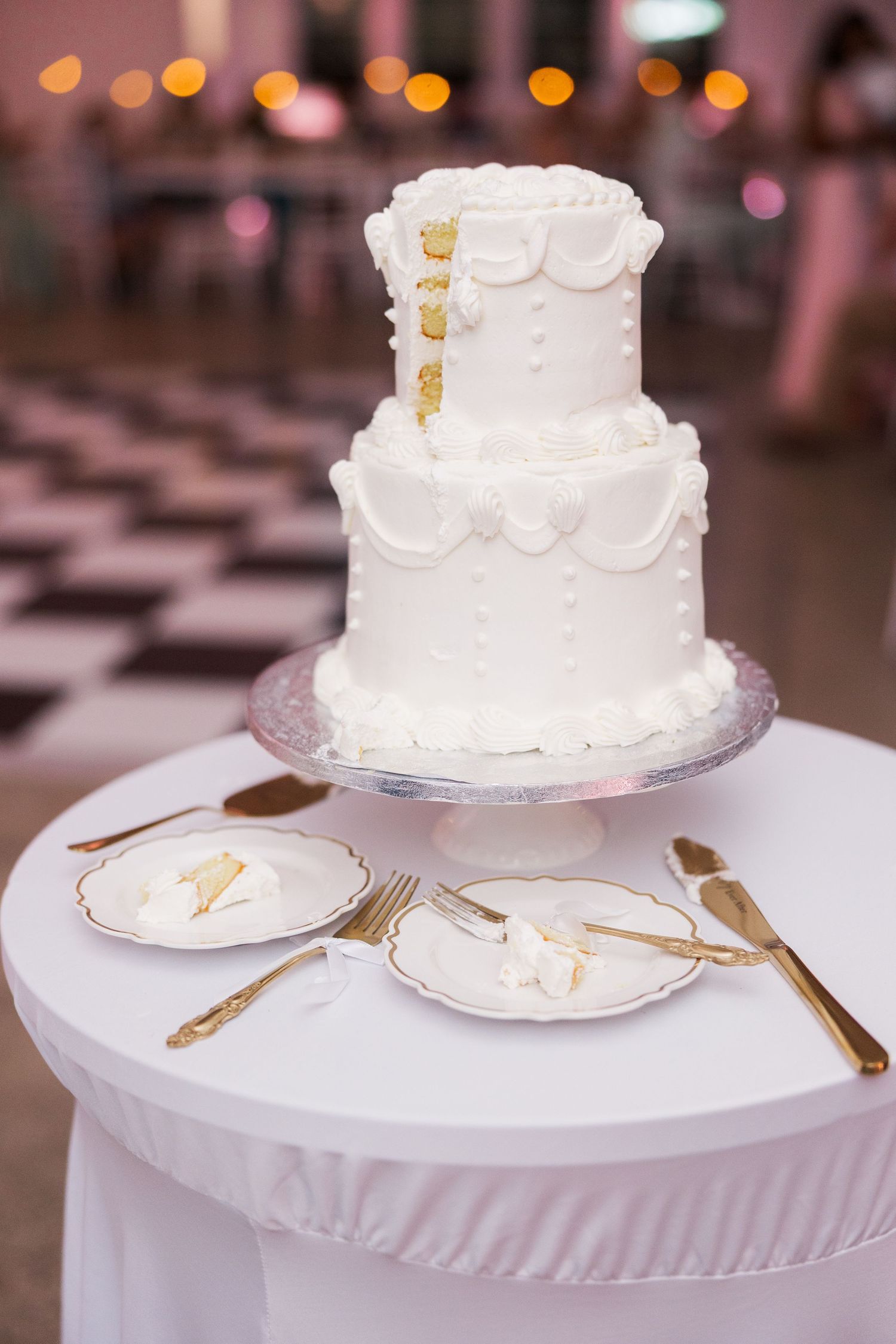 An elegant white tiered wedding cake displayed on a table with gold utensils and soft lighting.