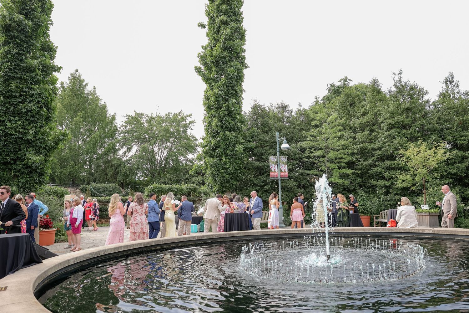 Wedding party and guests gathered around outdoor fountain feature in landscaped garden.