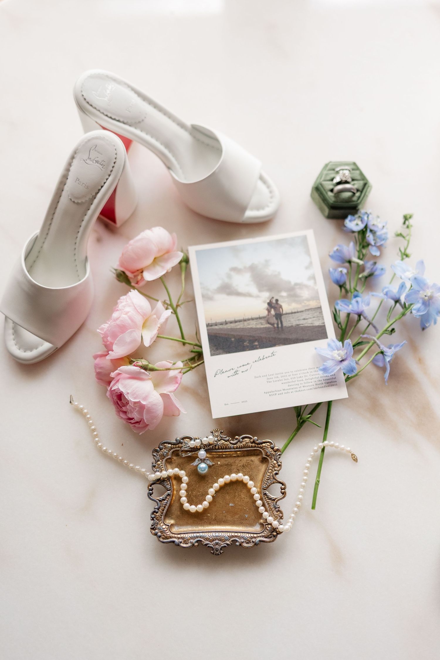 White bridal shoes and pearl jewelry displayed with pink roses and blue forget-me-nots on a light surface.