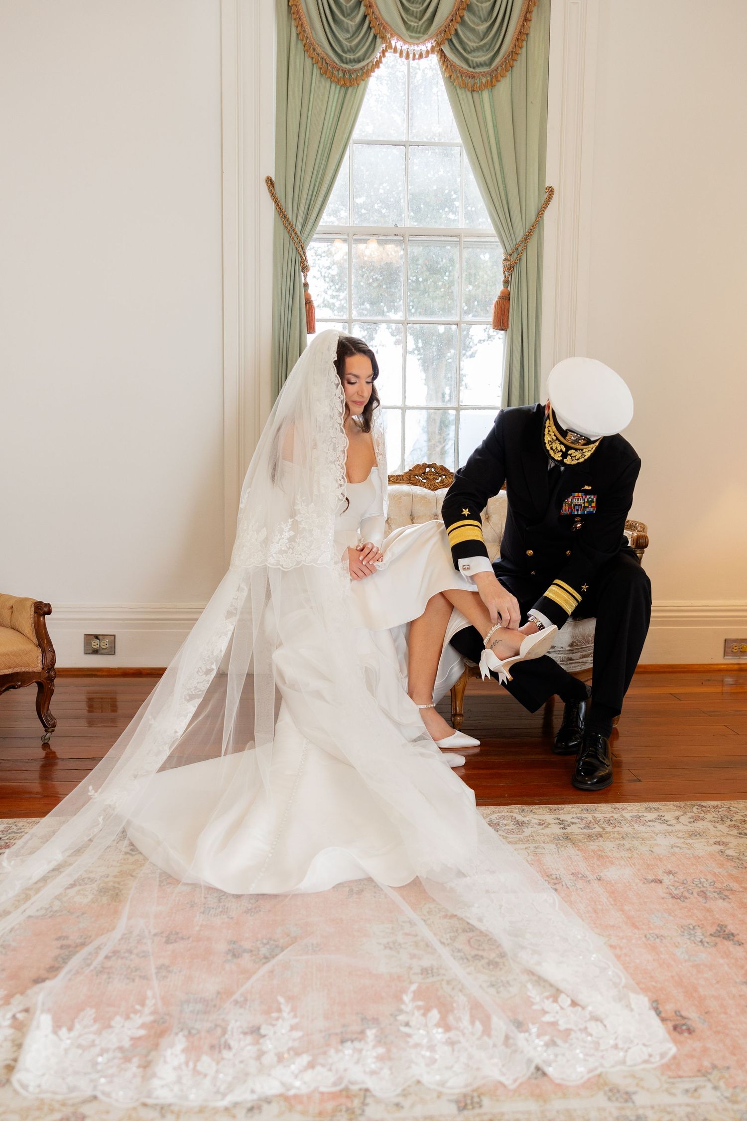 A bride in a flowing white wedding gown with a long lace train poses on an elegant floral carpet in an indoor setting.