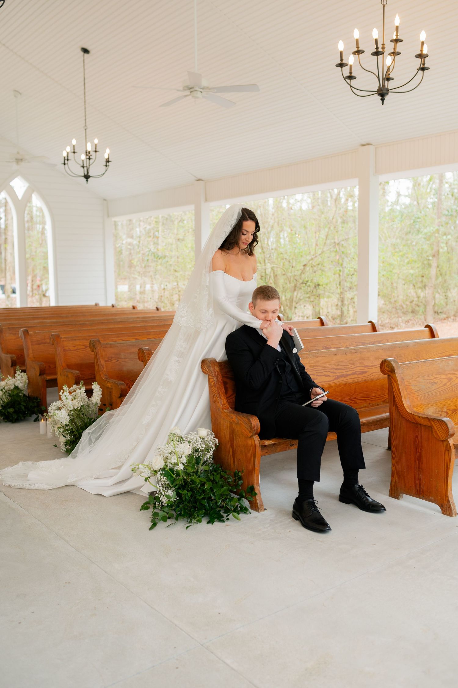 Wedding couple poses artistically in bright chapel with wooden pews and greenery decorations.