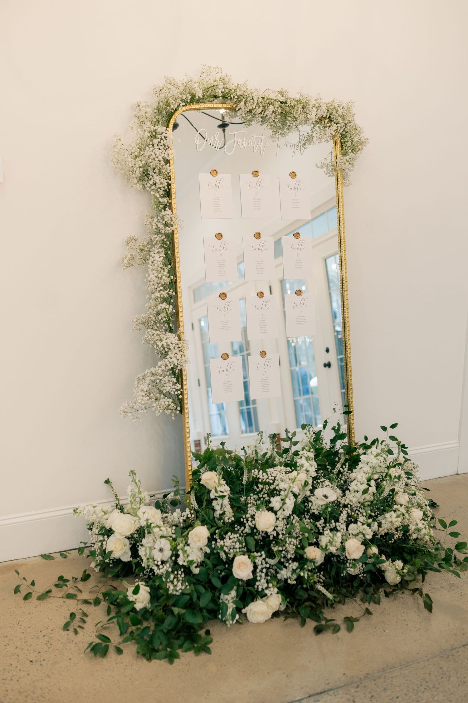 White floral seating chart display with greenery and roses arranged along the bottom.