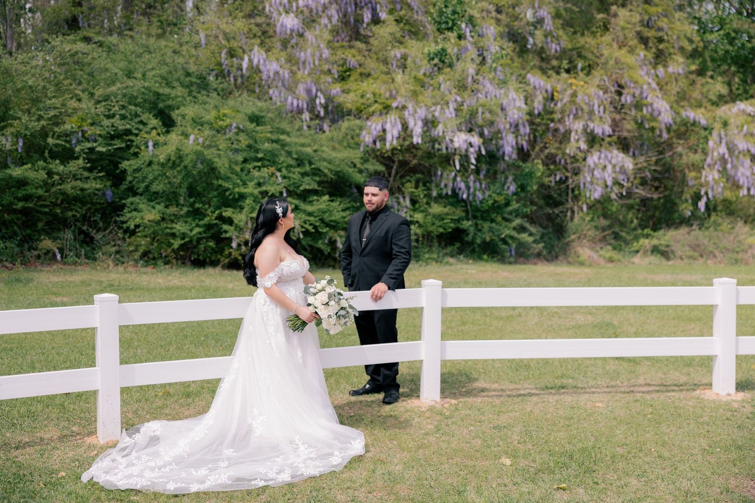 Wedding party poses along white fence with wisteria and greenery in background.