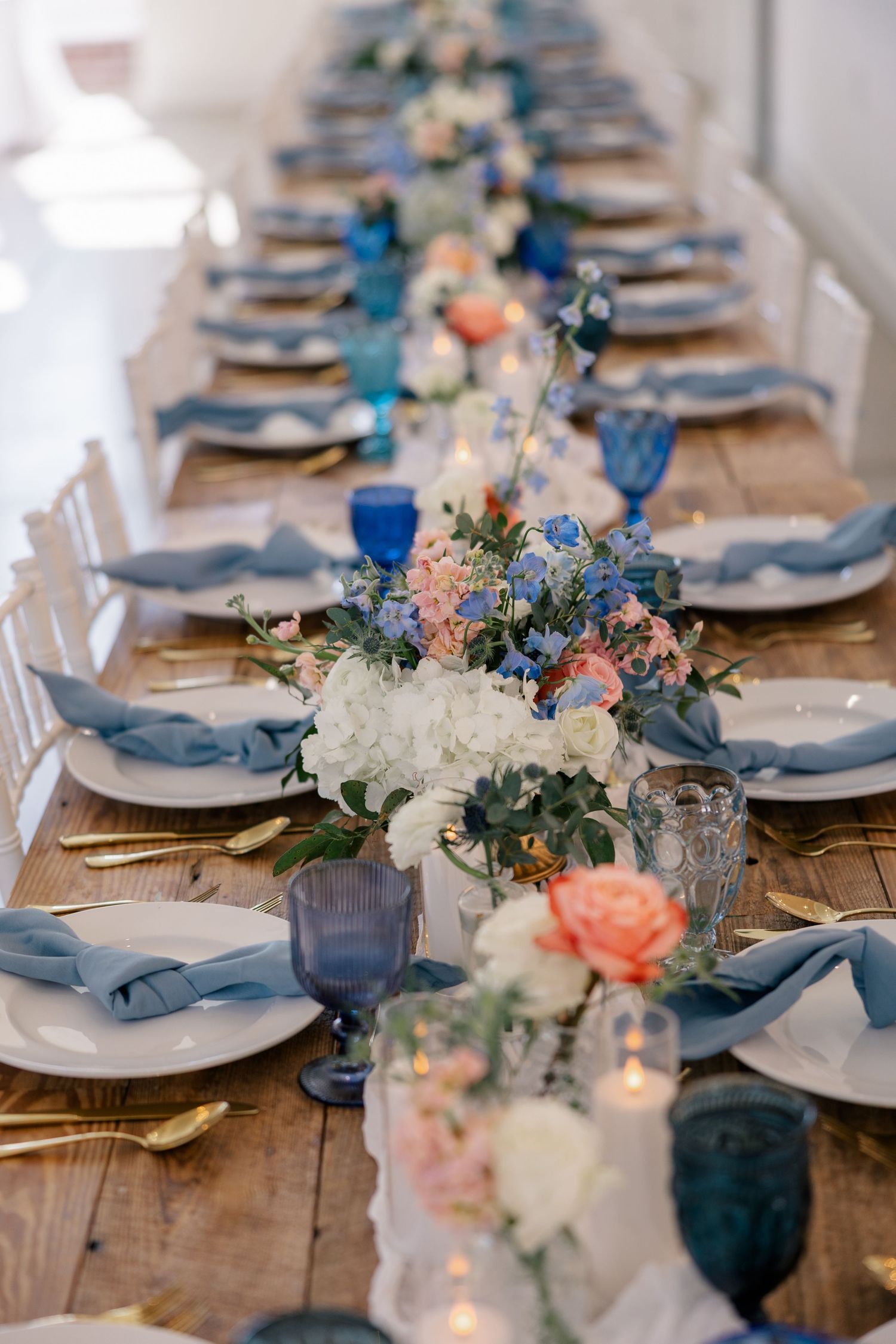 Elegant wedding table setting with blue glassware, white plates, and peach floral centerpieces on rustic wooden table.