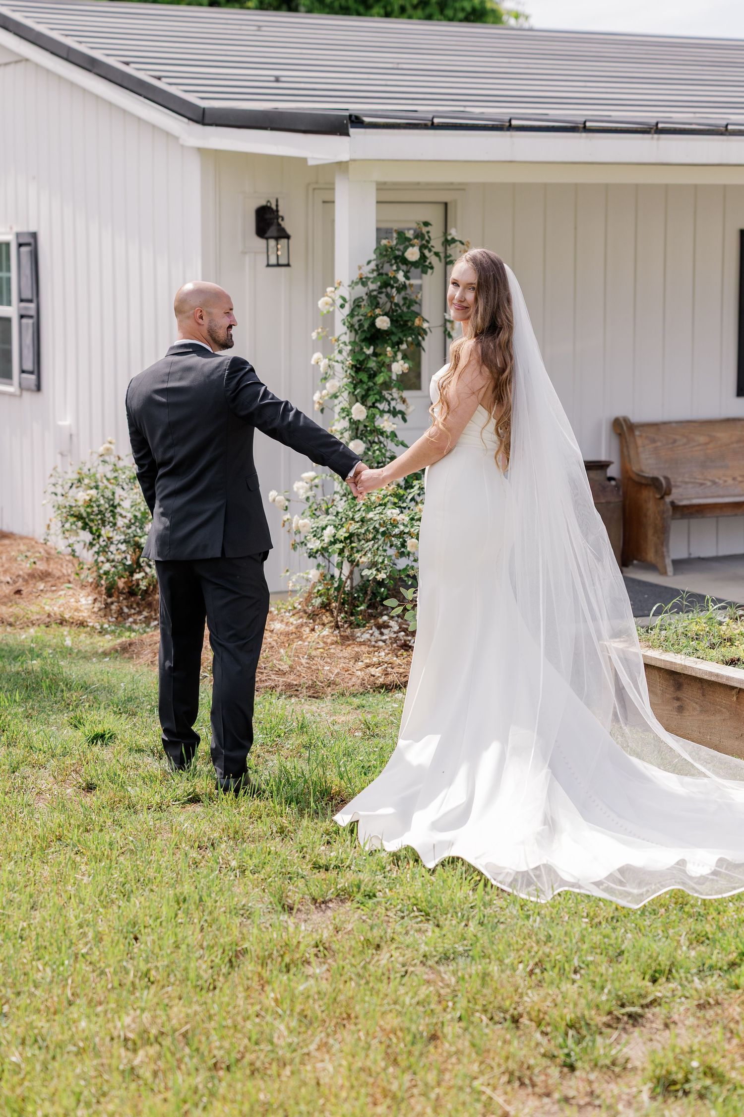 Bride in flowing white dress walking with someone across a grassy lawn outside a white chapel.