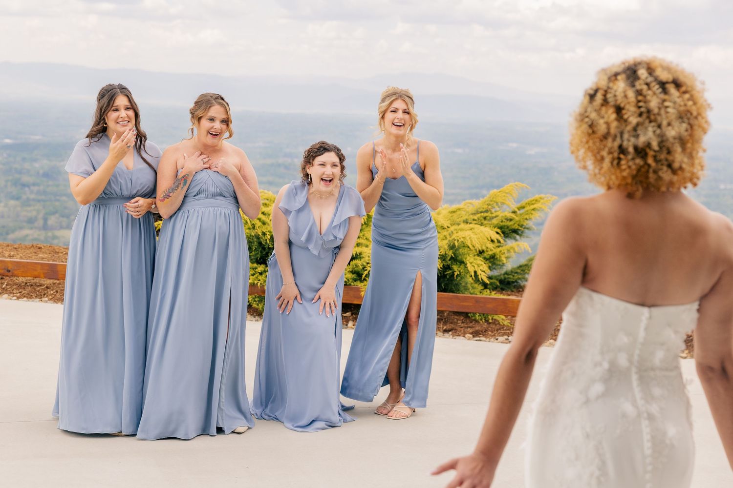 Bridesmaids in blue gowns react with joy seeing bride in white wedding dress at outdoor venue.