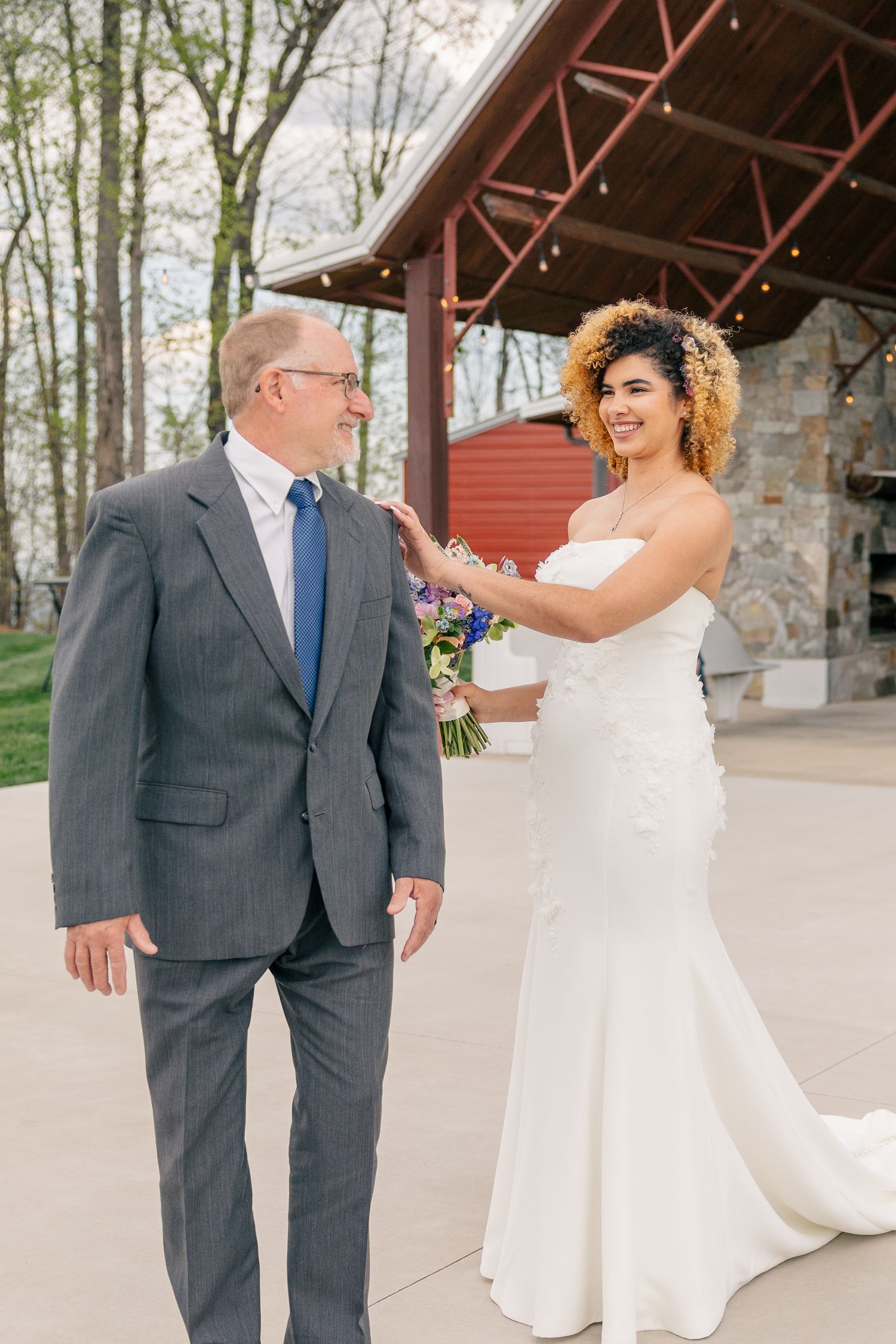 Bride in white gown holds bouquet, touching groom's shoulder in gray suit at outdoor wedding venue.