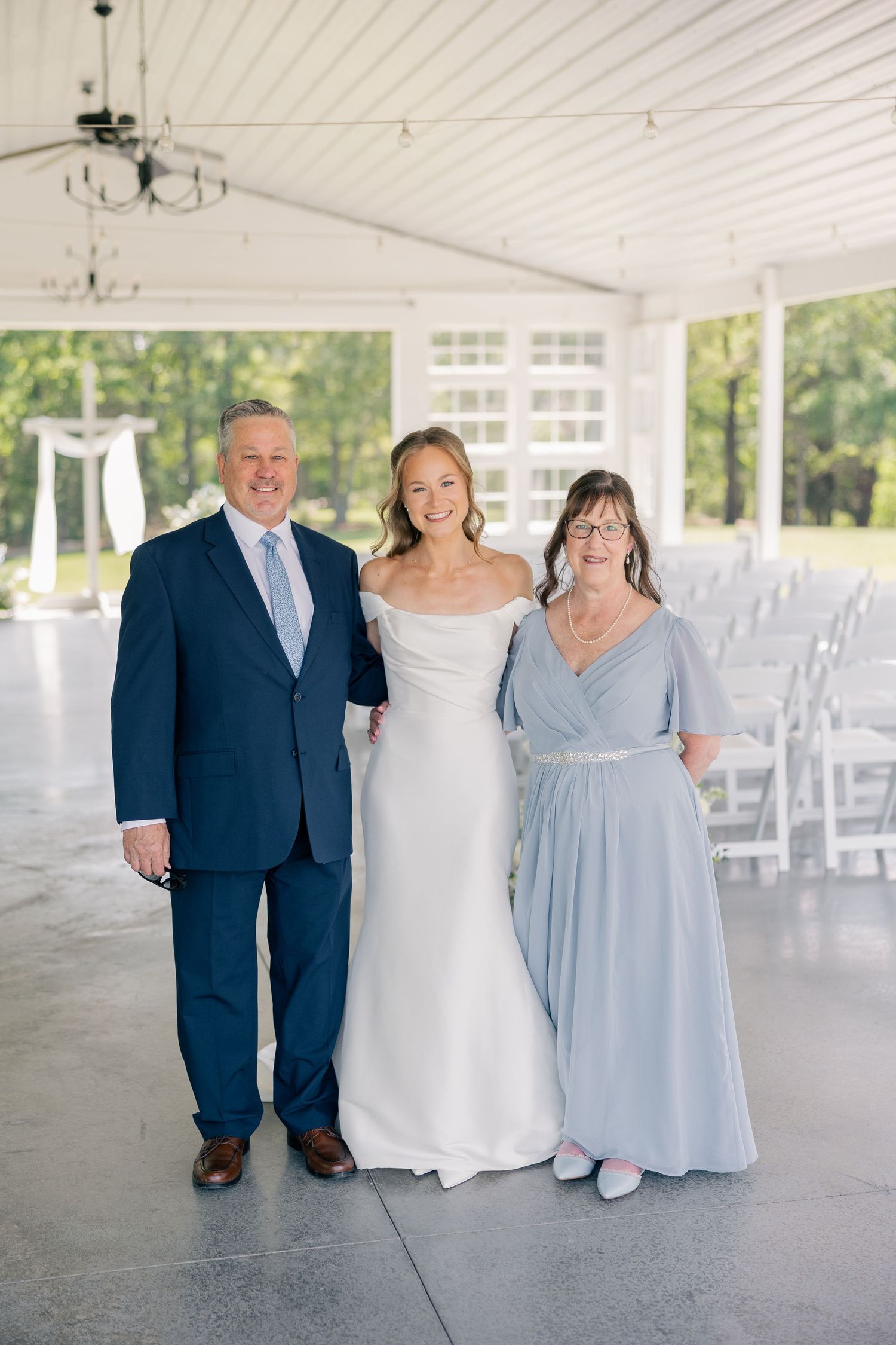 Bride in off-shoulder white gown poses with parents in blue attire at elegant outdoor wedding venue.