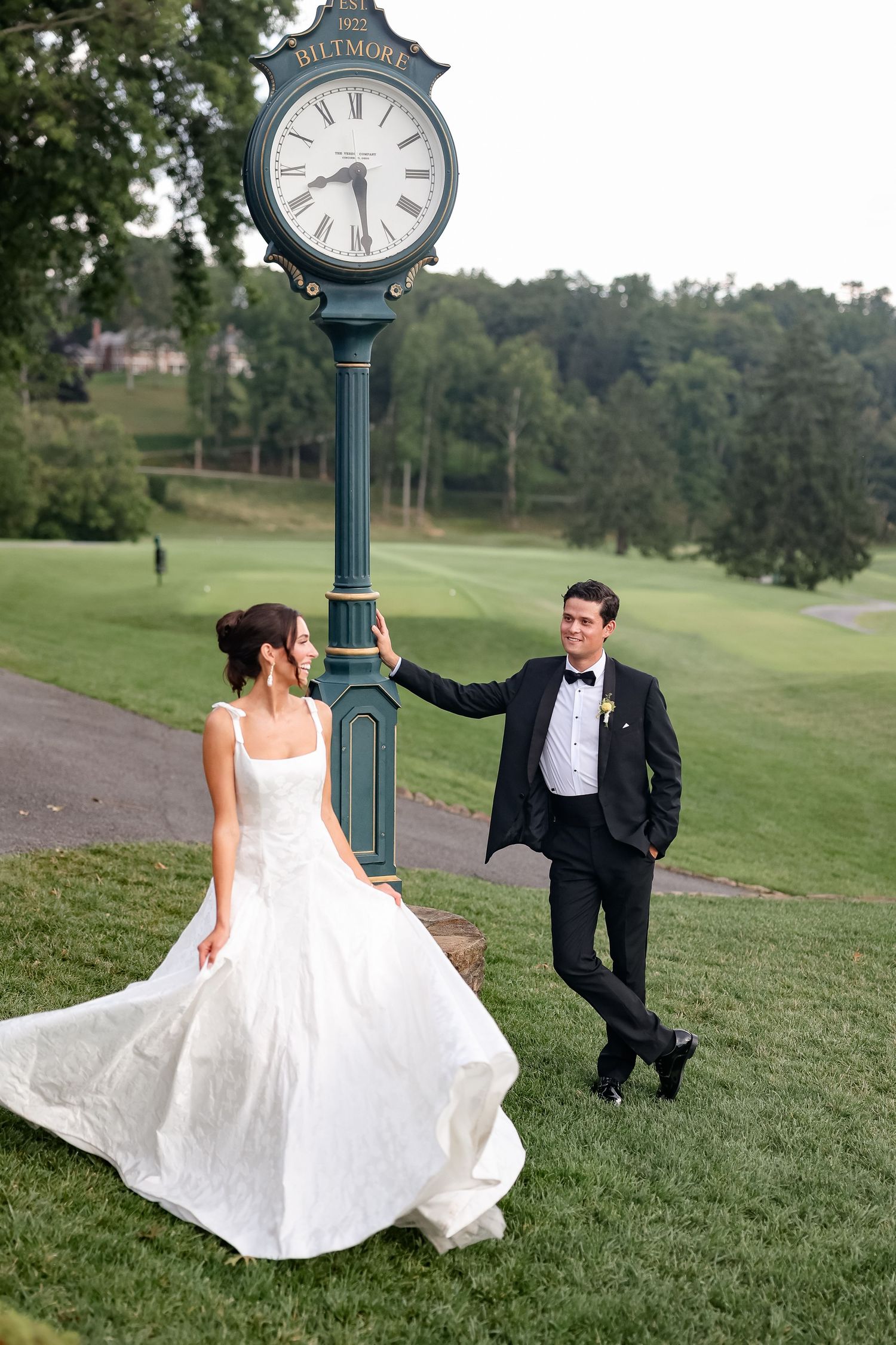 Bride and groom pose by a Biltmore clock tower on a lush green golf course on their wedding day.