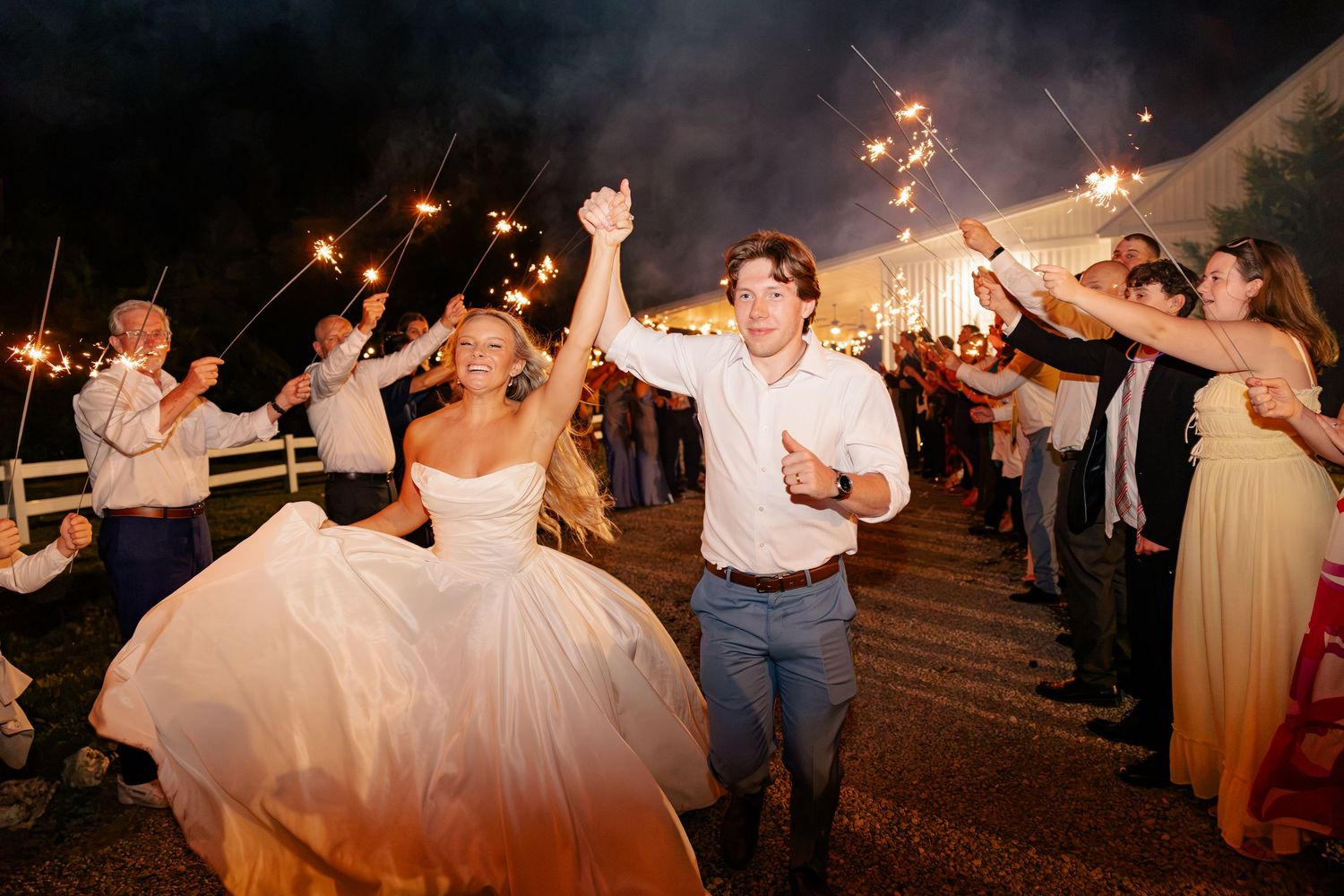 Bride and groom exit through sparkler tunnel at night, guests celebrating with raised sparklers at wedding reception.