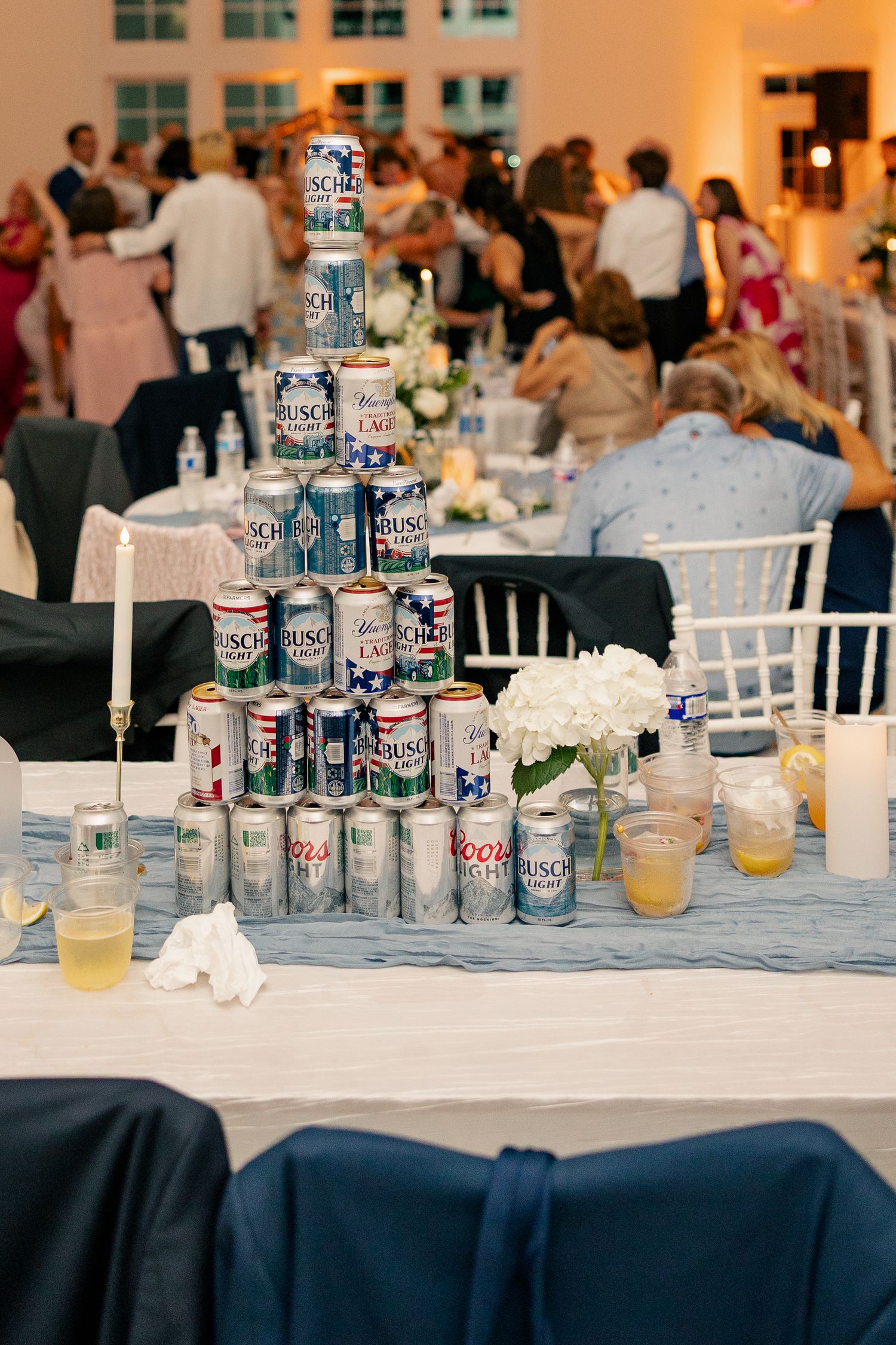 Pyramid of Busch Light beer cans on blue linen table at lively wedding reception.