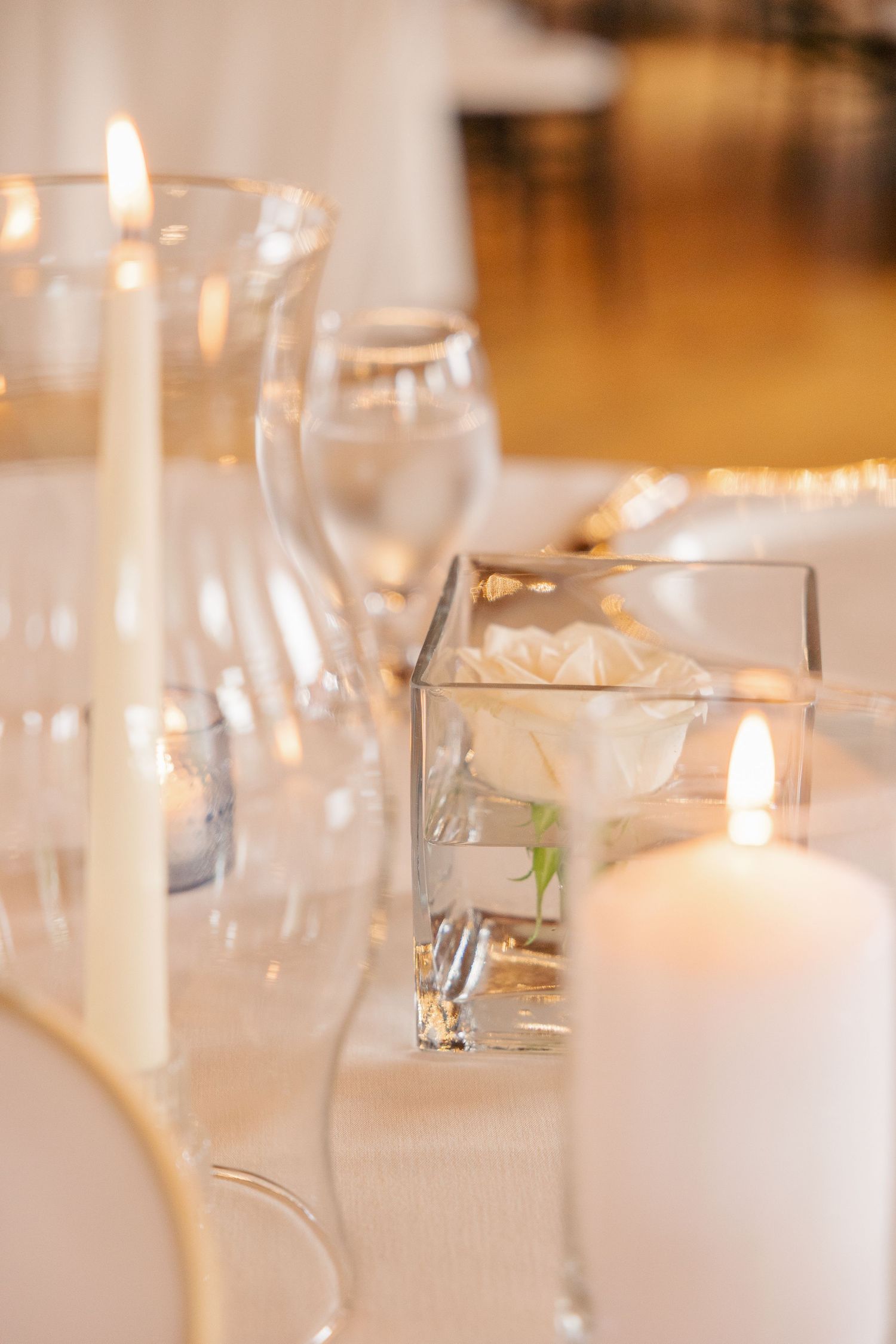 Elegant wedding table setting with white candles, glass vase with rose, and crystal glassware.