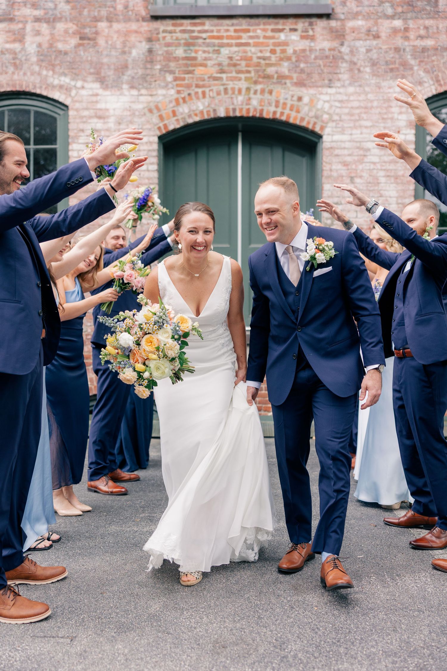 Happy newlyweds walk through arch of raised hands formed by wedding party in navy suits.