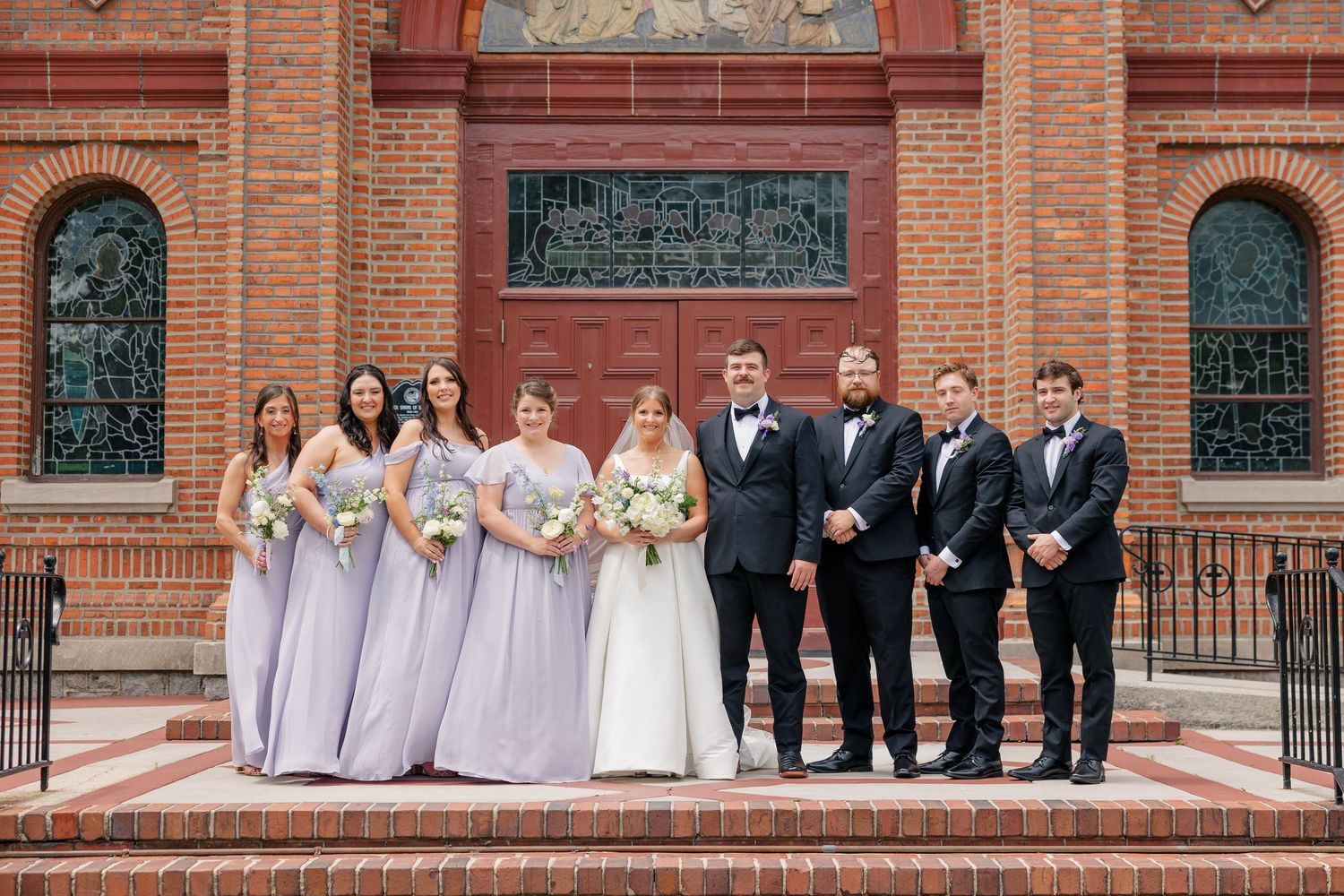Wedding party poses in front of brick church, bridesmaids in lavender gowns, groomsmen in black tuxedos.
