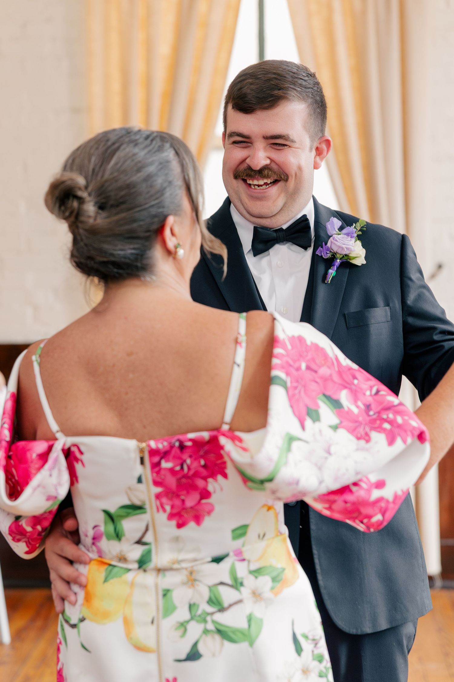 Groom in tuxedo laughing with a guest in floral dress at wedding reception.