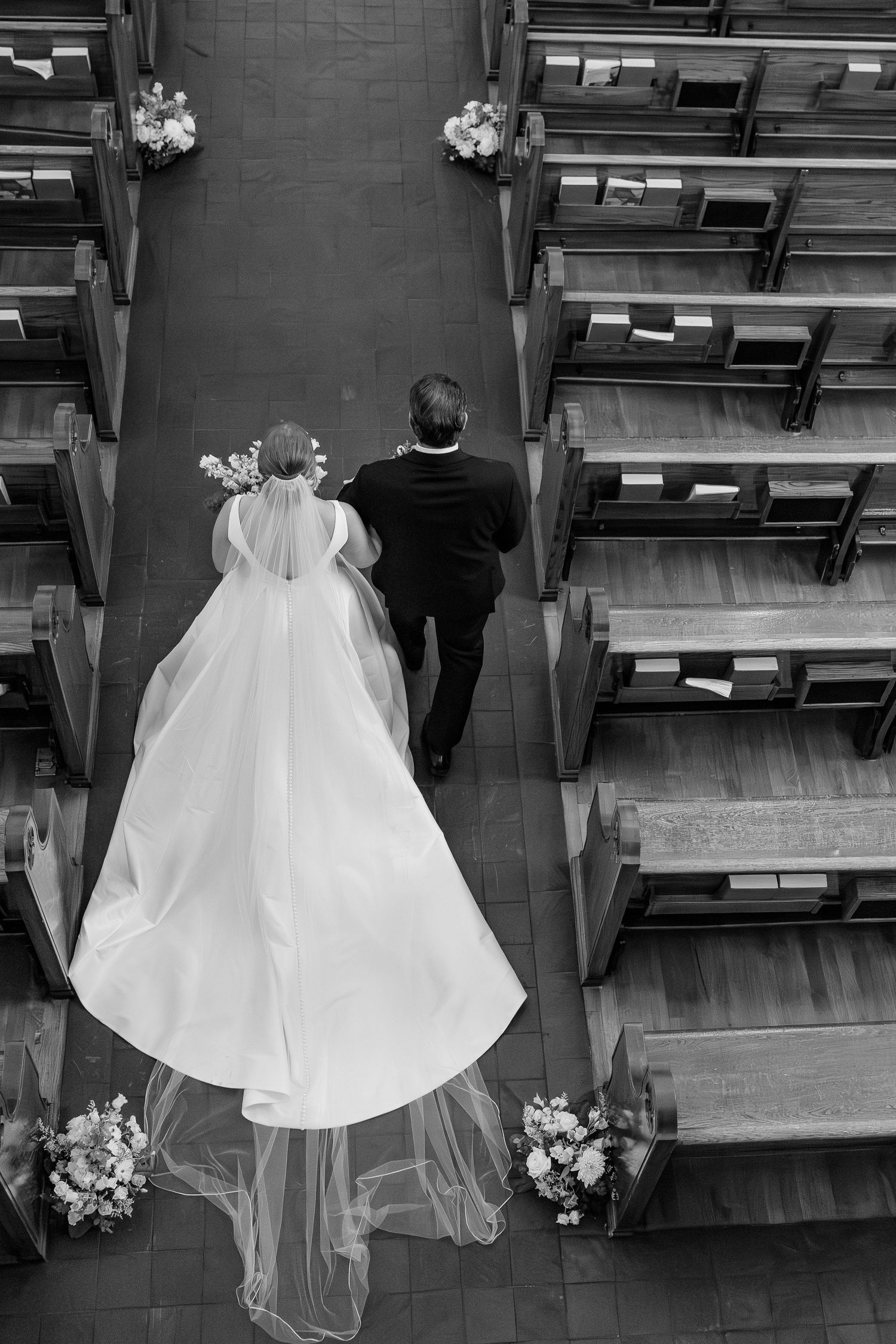 Aerial black and white photo of bride and groom walking down church aisle with flowing cathedral veil trailing behind.