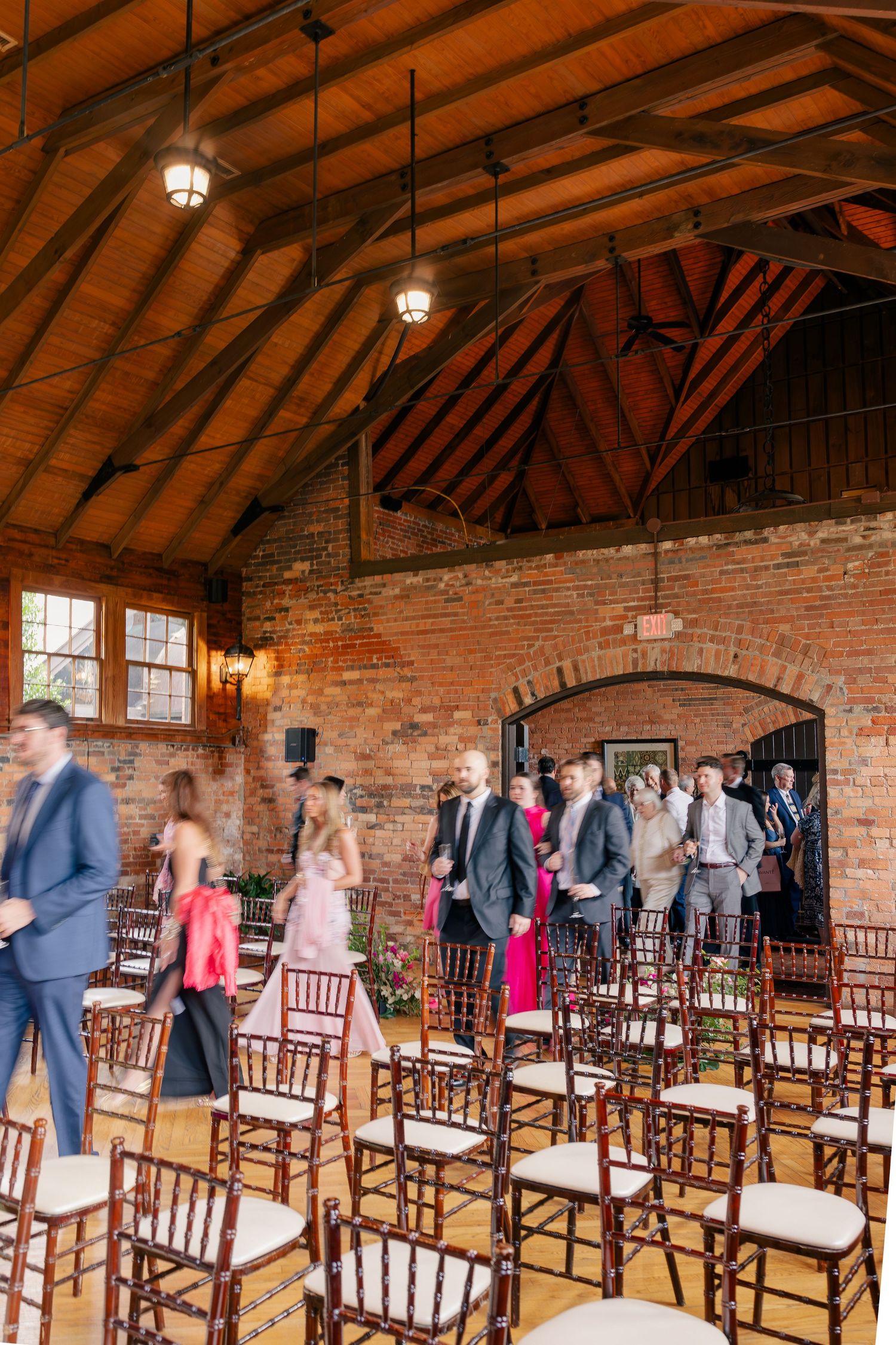 Guests arrive at a rustic brick wedding venue with wooden cathedral ceiling and chiavari chairs.