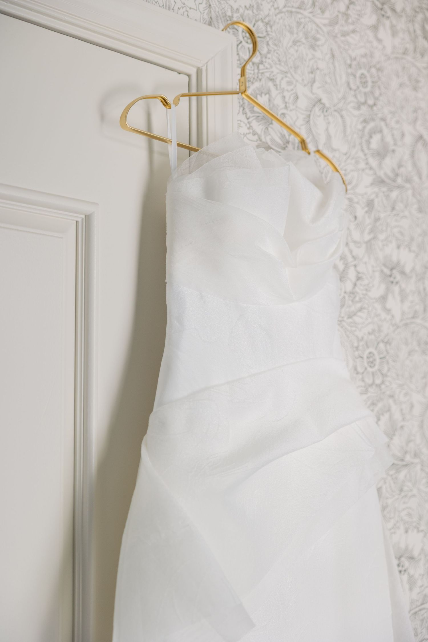Elegant white strapless wedding gown hanging on gold hanger against white door and floral wallpaper.