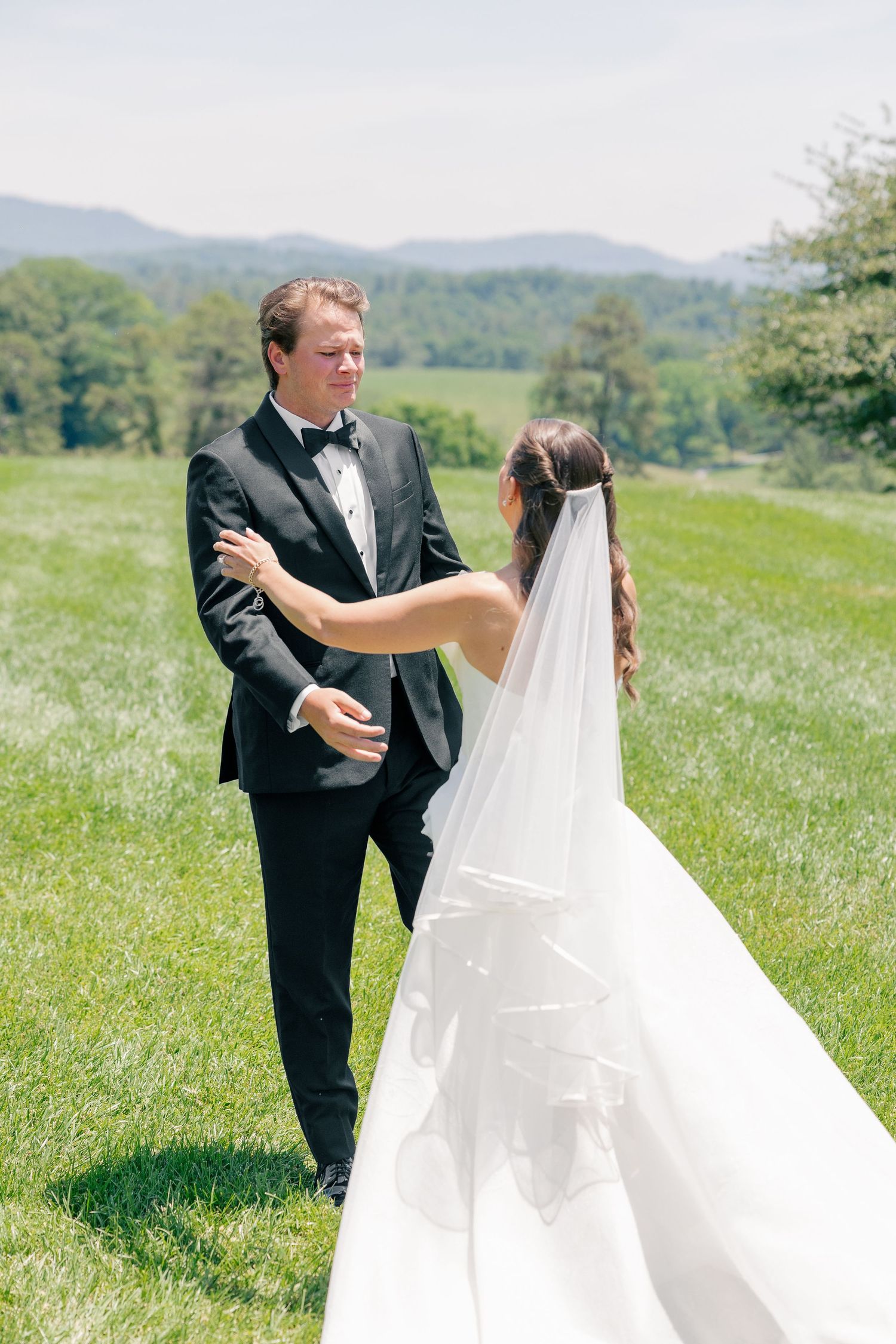 Groom seeing bride in white gown and veil for first time during outdoor first look on green hillside.