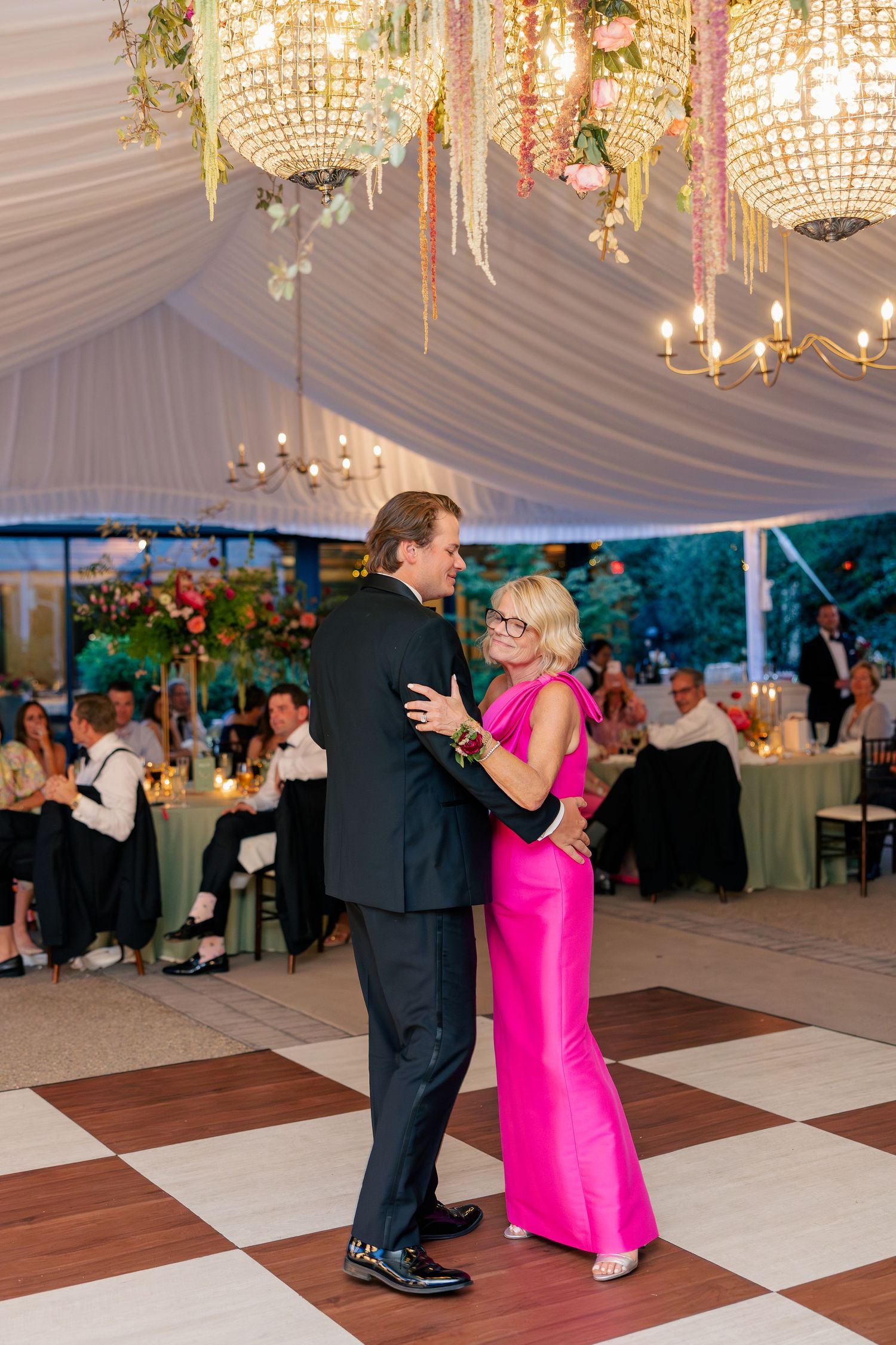 Groom dances with mother wearing a vibrant fuchsia gown under crystal chandeliers at wedding reception.