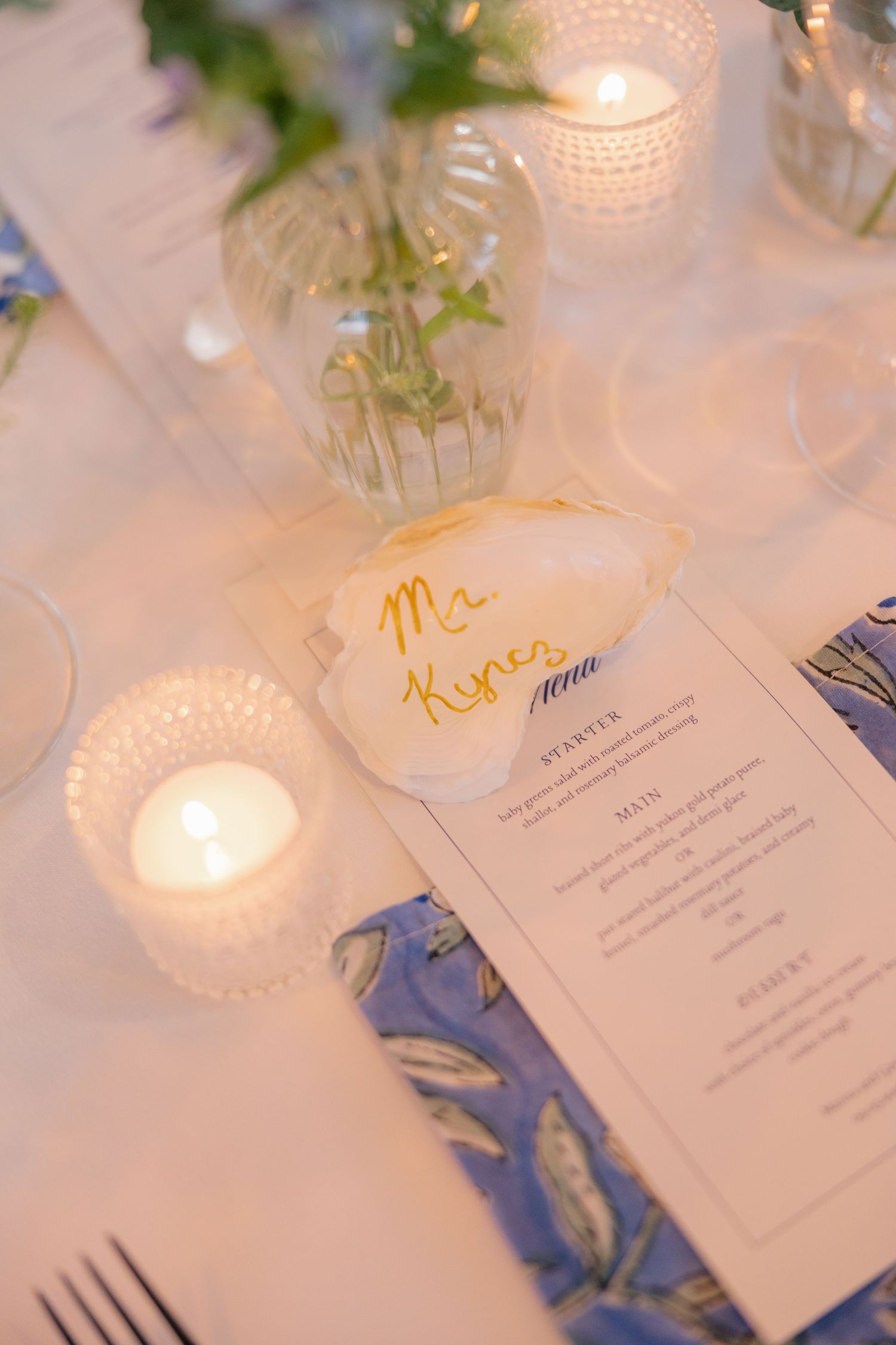 Elegant wedding place setting with handwritten name card, blue floral napkin, menu, and candles.