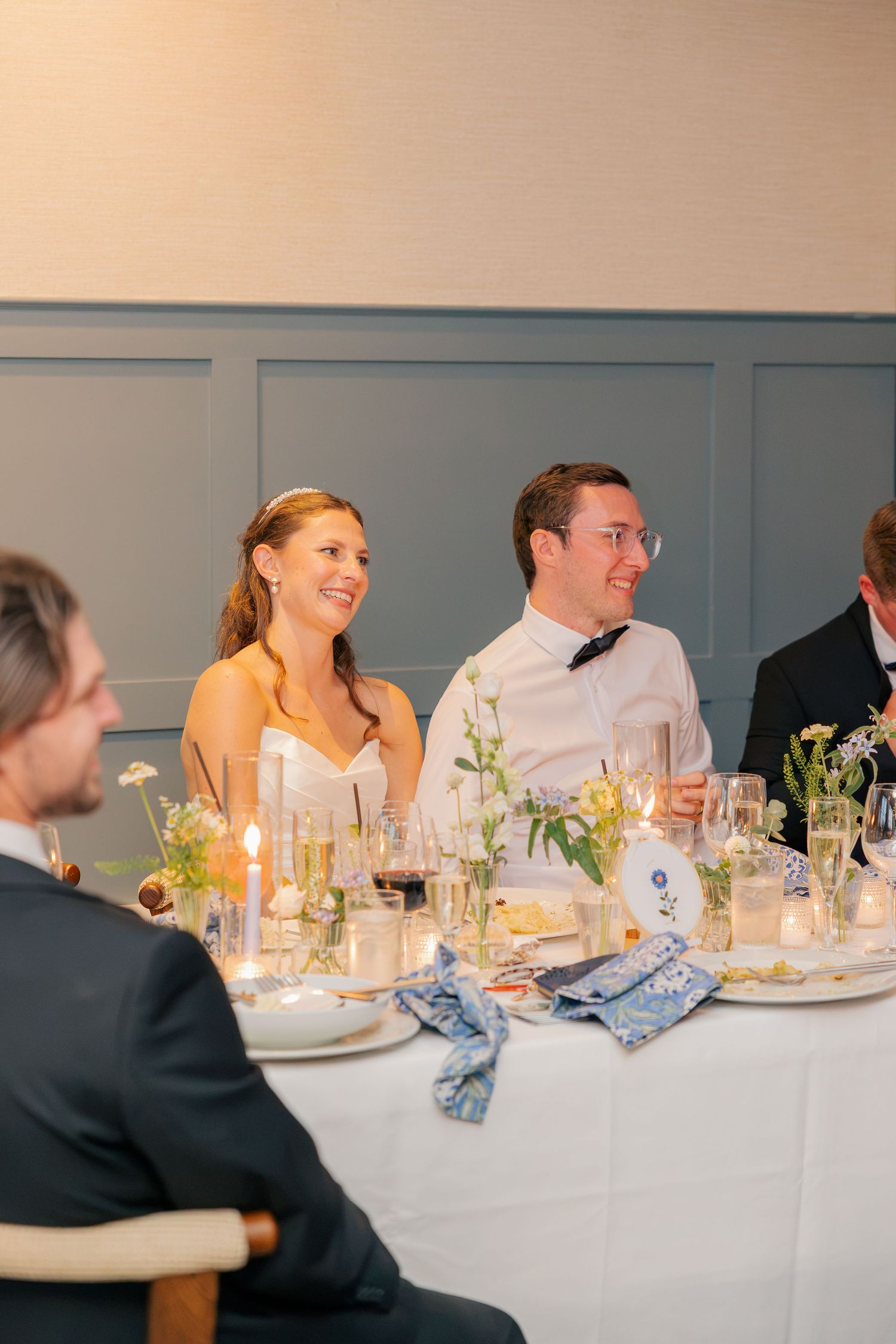 Bride and groom smiling at wedding reception dinner table with candles and floral arrangements.