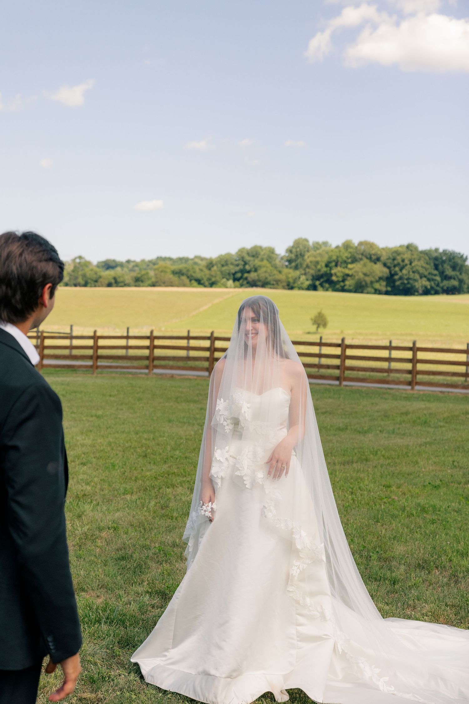 Bride in cathedral veil and floral gown walks toward groom on green farm field under blue sky.