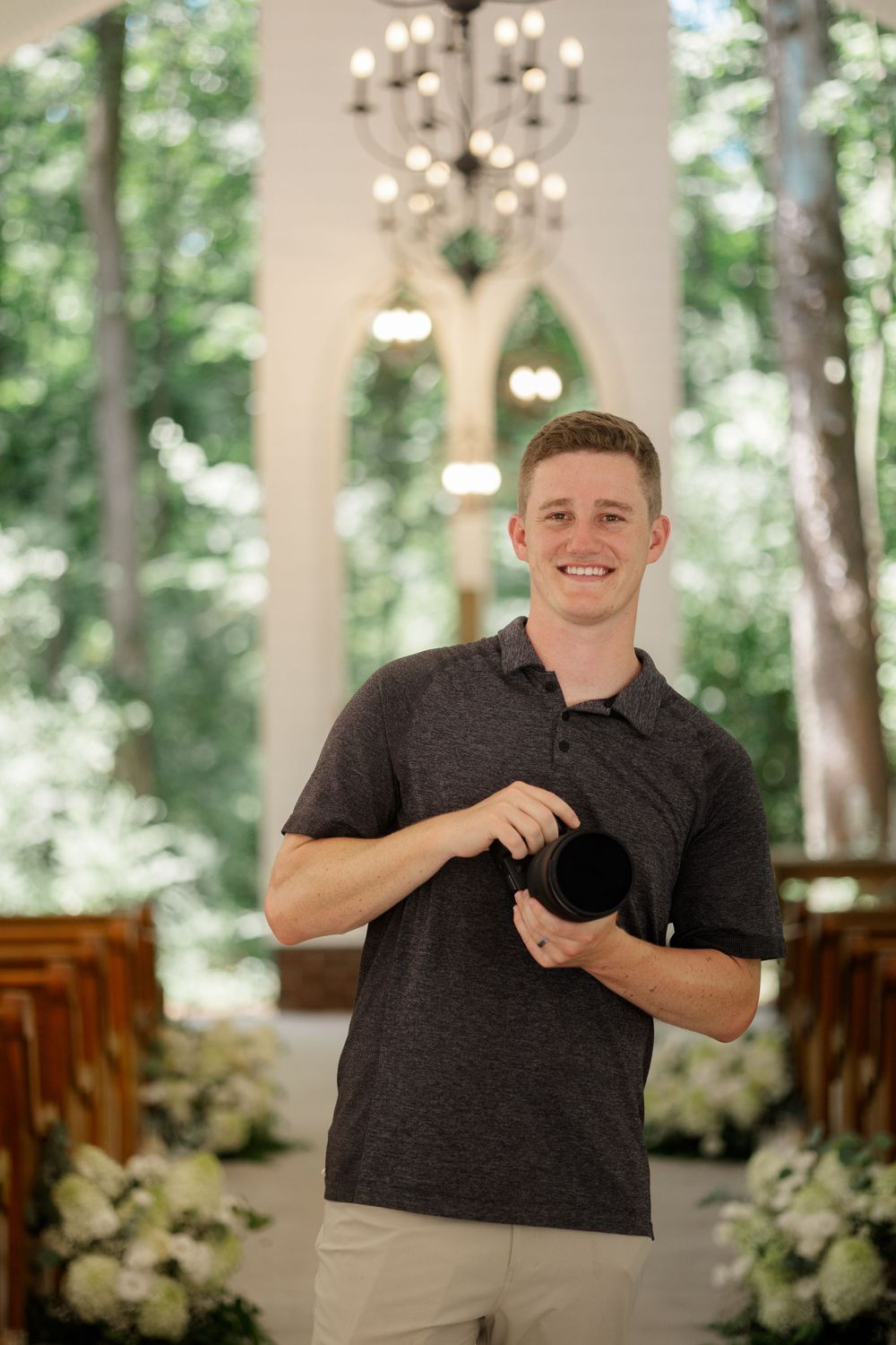 Wedding photographer in dark polo shirt holding camera in chapel setting with chandeliers.