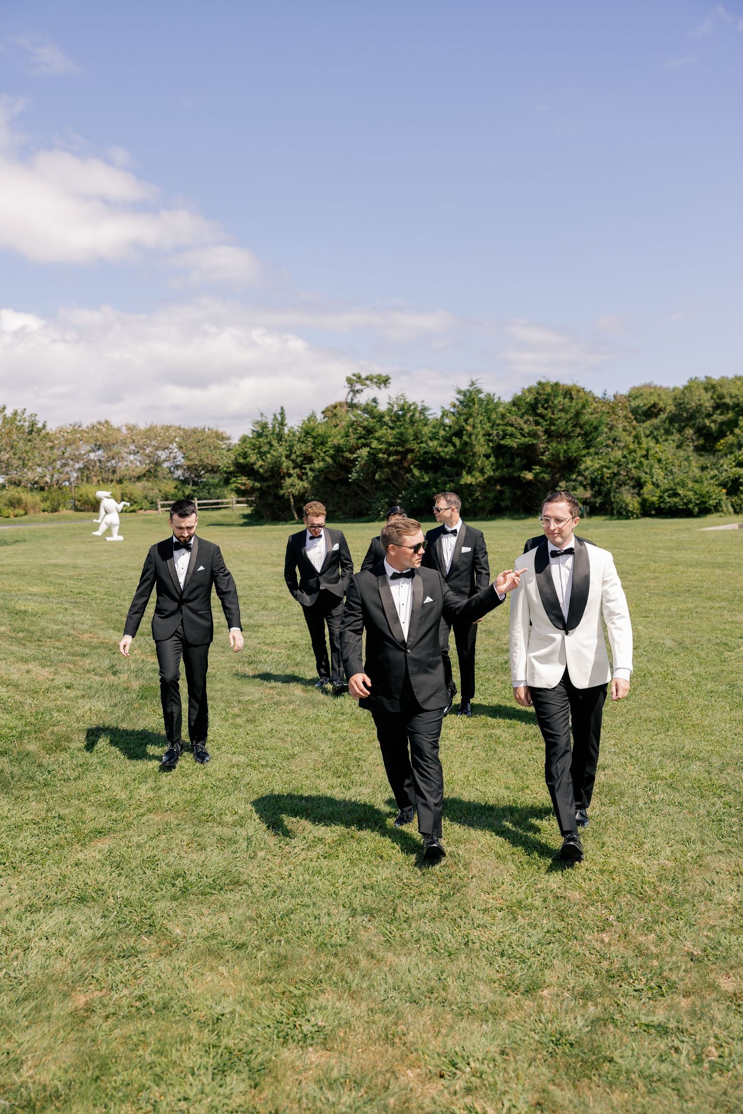 Groom in white jacket walks with groomsmen in black tuxedos across green lawn outdoors.