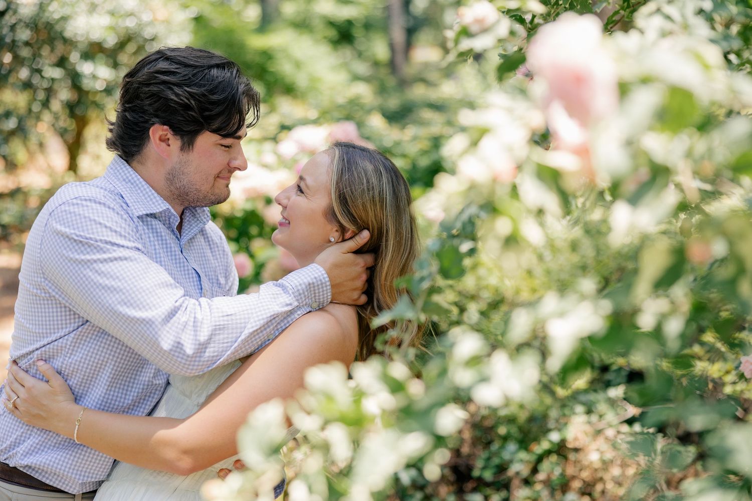 Couple embracing and smiling at each other surrounded by lush green foliage and pink flowers outdoors.