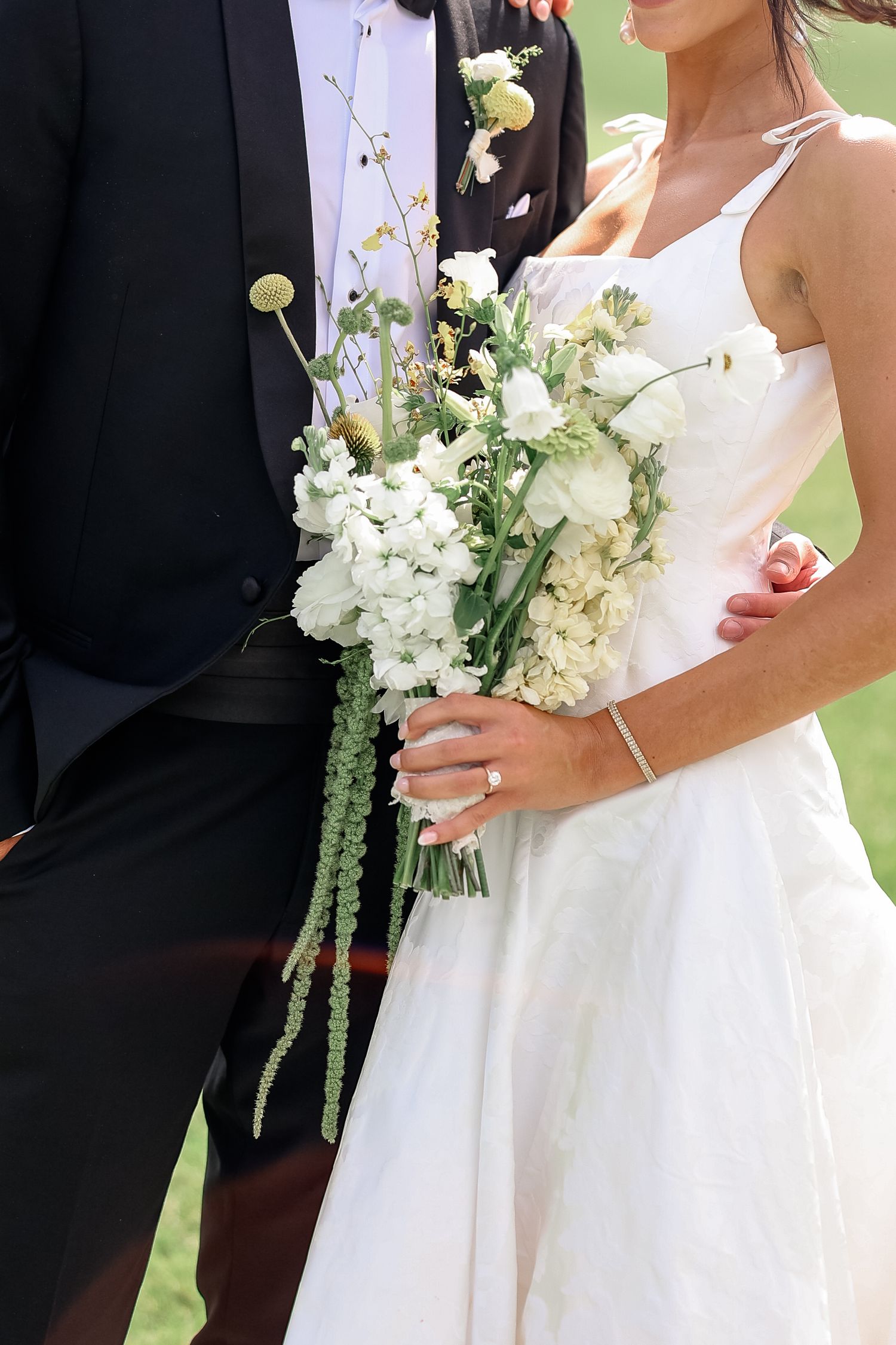 Bride and groom standing close together holding white and green wedding bouquet outdoors.