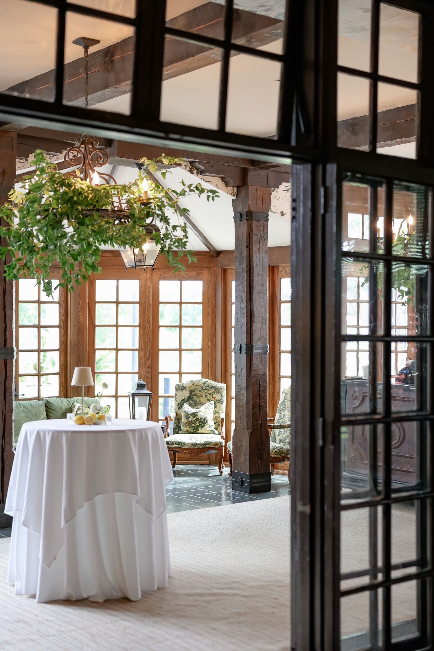 Elegant sunroom with white-clothed cocktail table, wood beams, French doors, and lush hanging greenery chandelier.