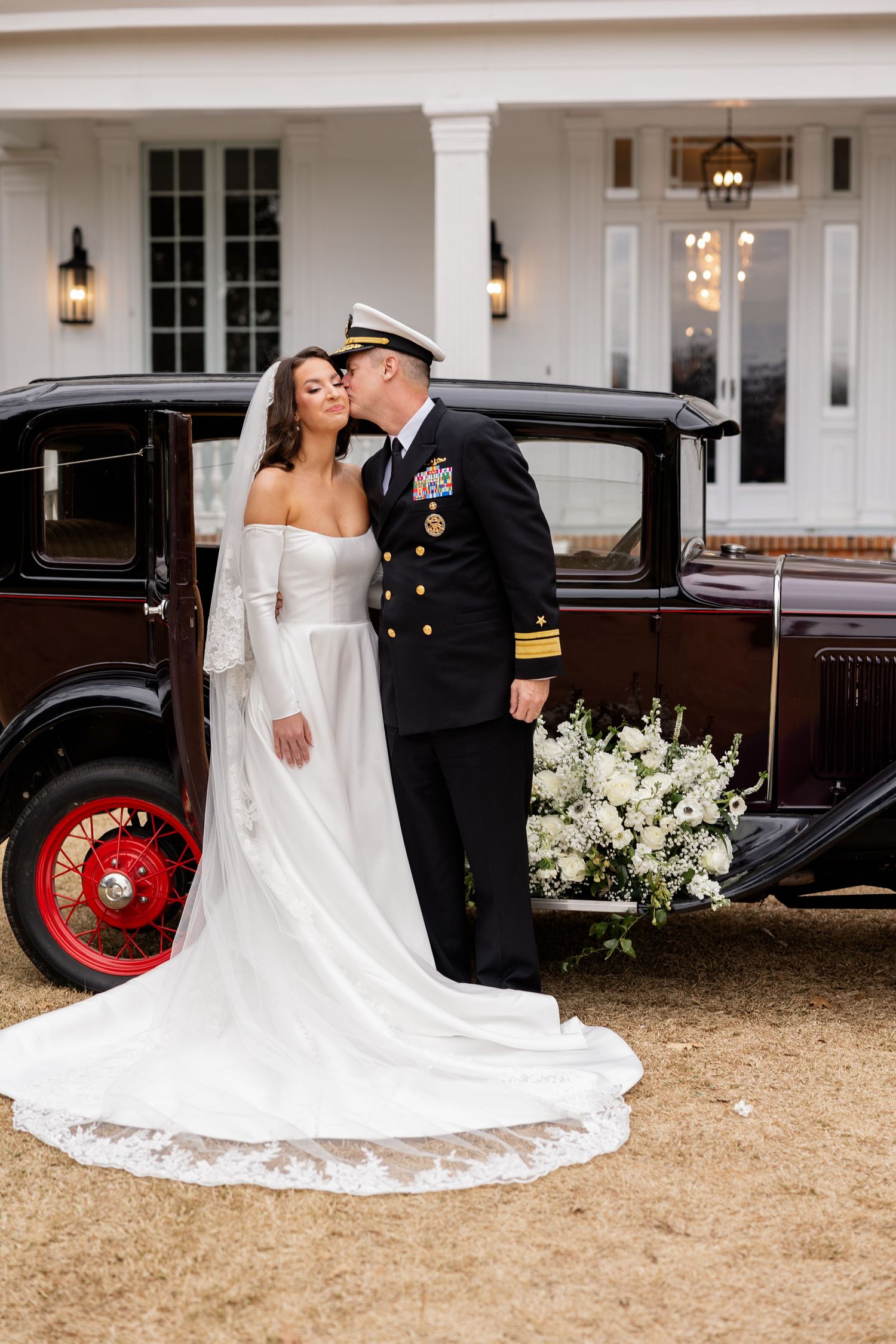 Military wedding couple poses romantically in front of a classic car and elegant white mansion.