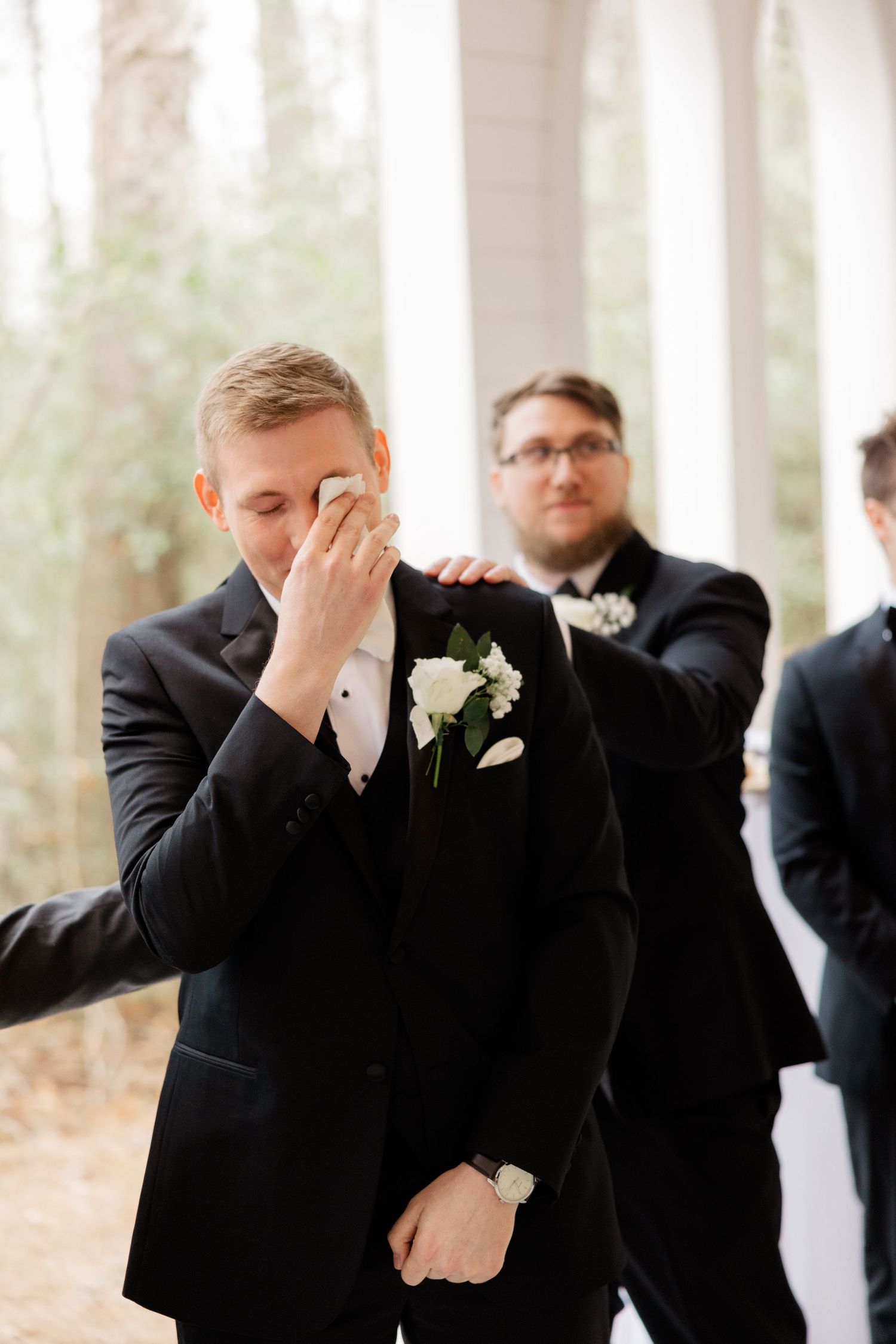 An emotional moment at a wedding ceremony shows a groom in a black suit wiping tears as another groomsman looks on.