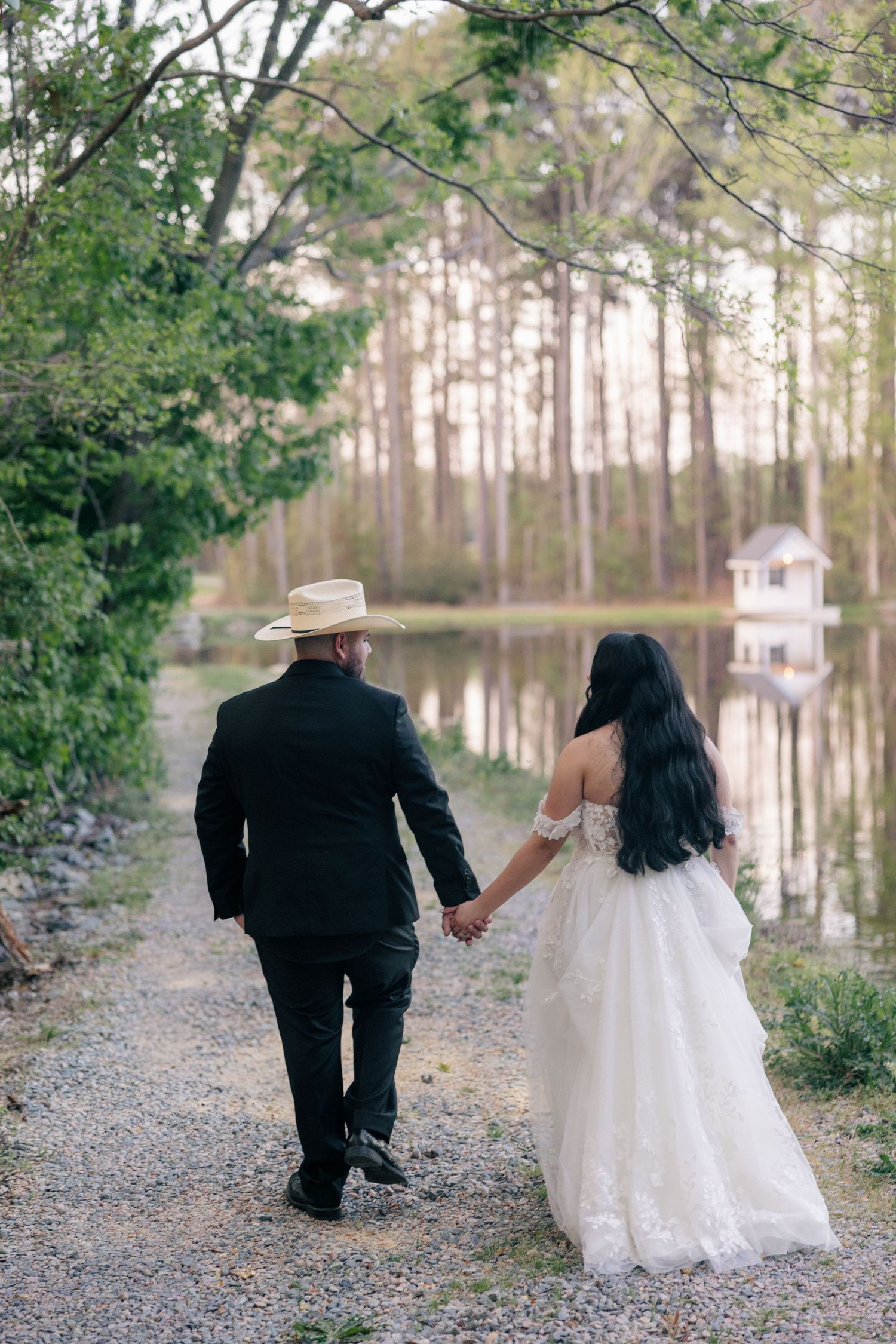 A couple walks hand in hand along a scenic path by a lake surrounded by pine trees.