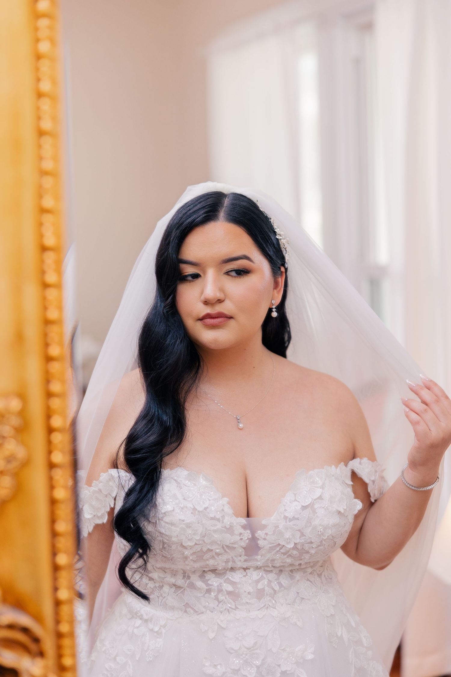 A bride in an off-shoulder lace wedding gown with long dark hair and veil poses near a gold-framed mirror.