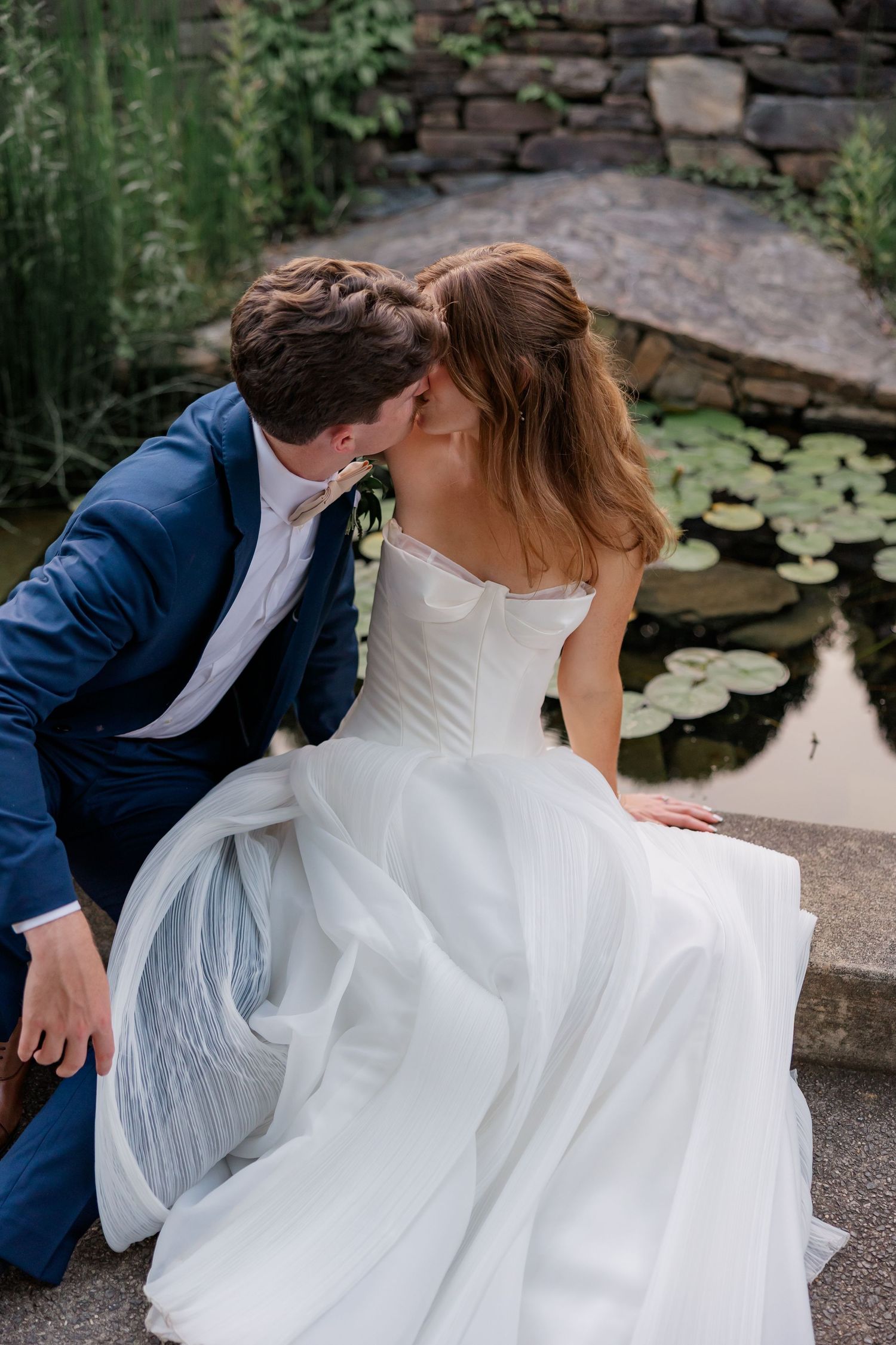 Wedding couple sitting by a lily pond with flowing white dress cascading over stone edge.