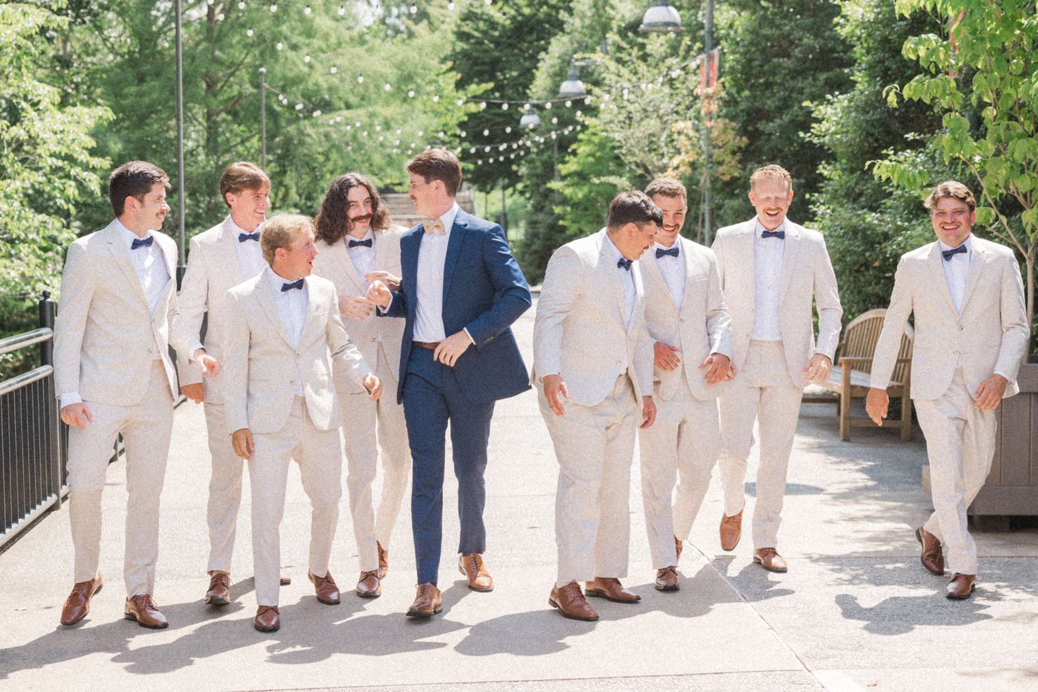 Wedding party members in light colored suits walk together in a line along an outdoor pathway.
