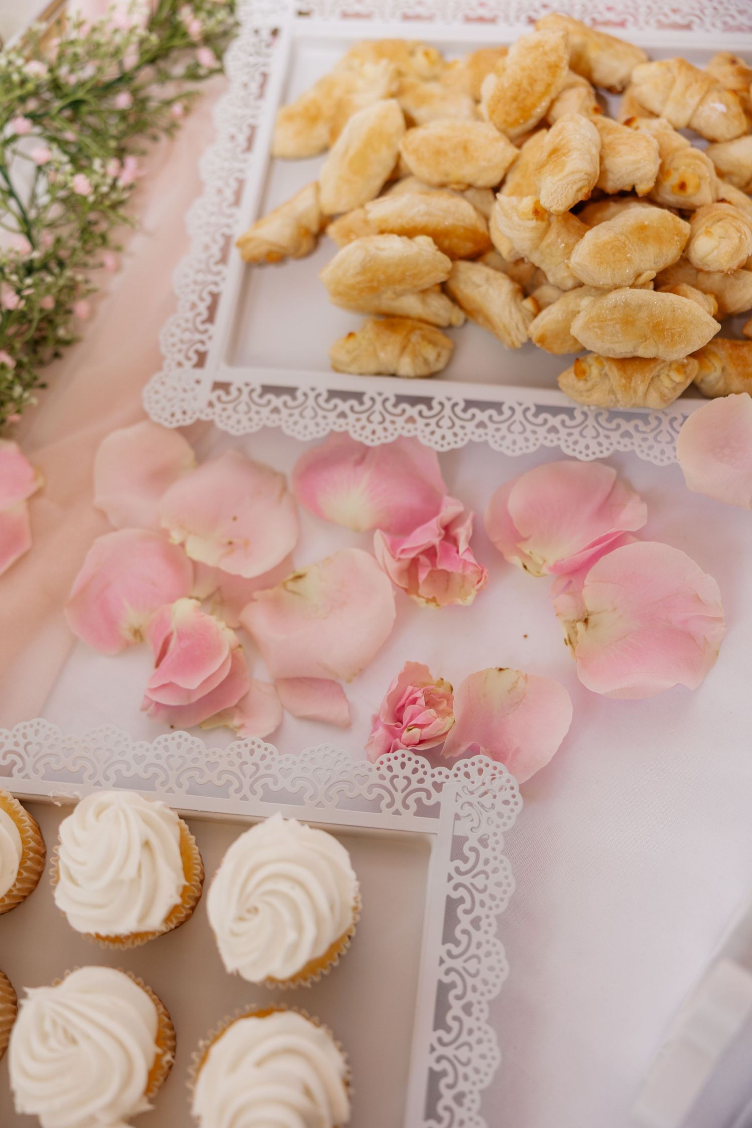 Wedding dessert display with mini croissants, cupcakes and pink rose petals on lace-trimmed trays.