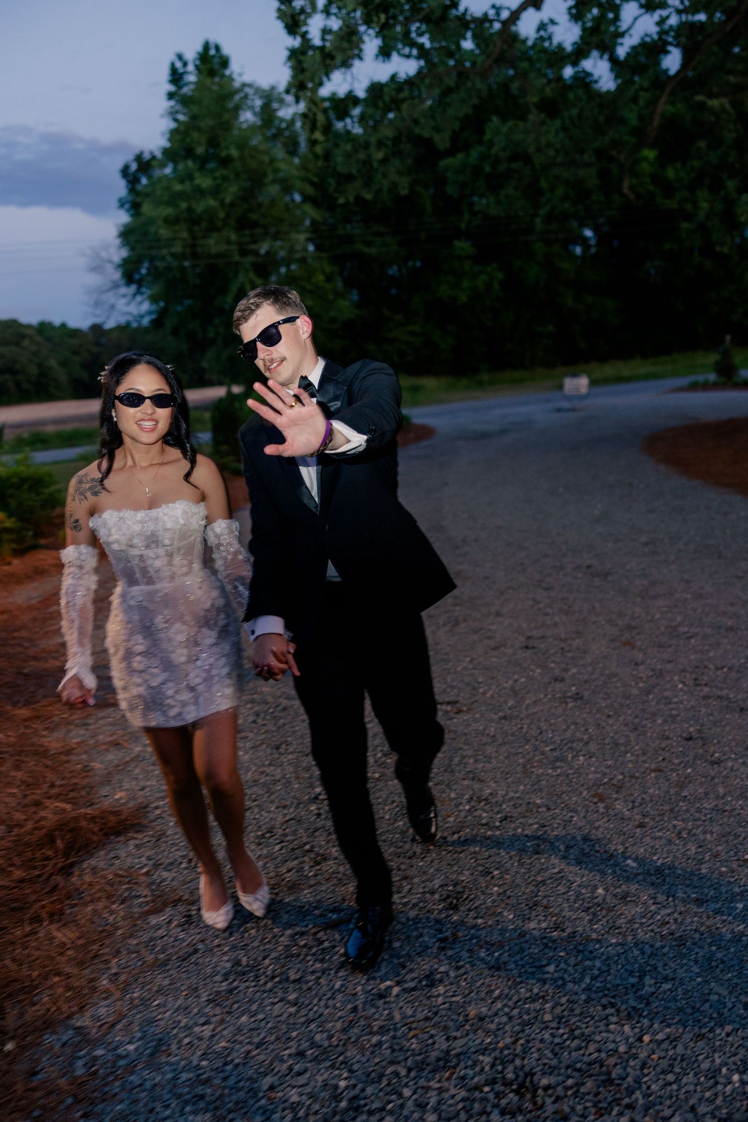 Playful couple in wedding attire posing with sunglasses on a gravel path at dusk.