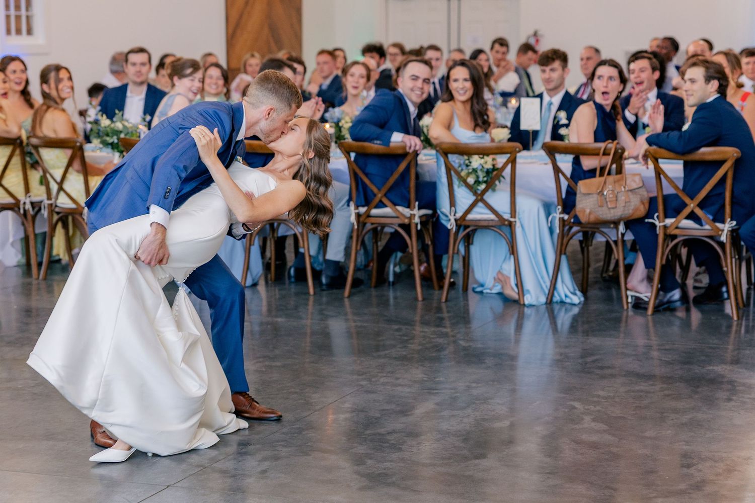 Groom dips bride during first dance at wedding reception while guests cheer and applaud.