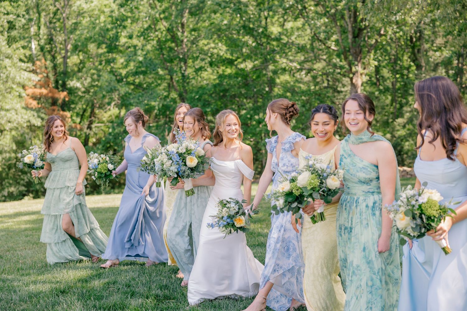 Bridesmaids in mismatched blue and green floral dresses walk together laughing while holding bouquets outdoors.