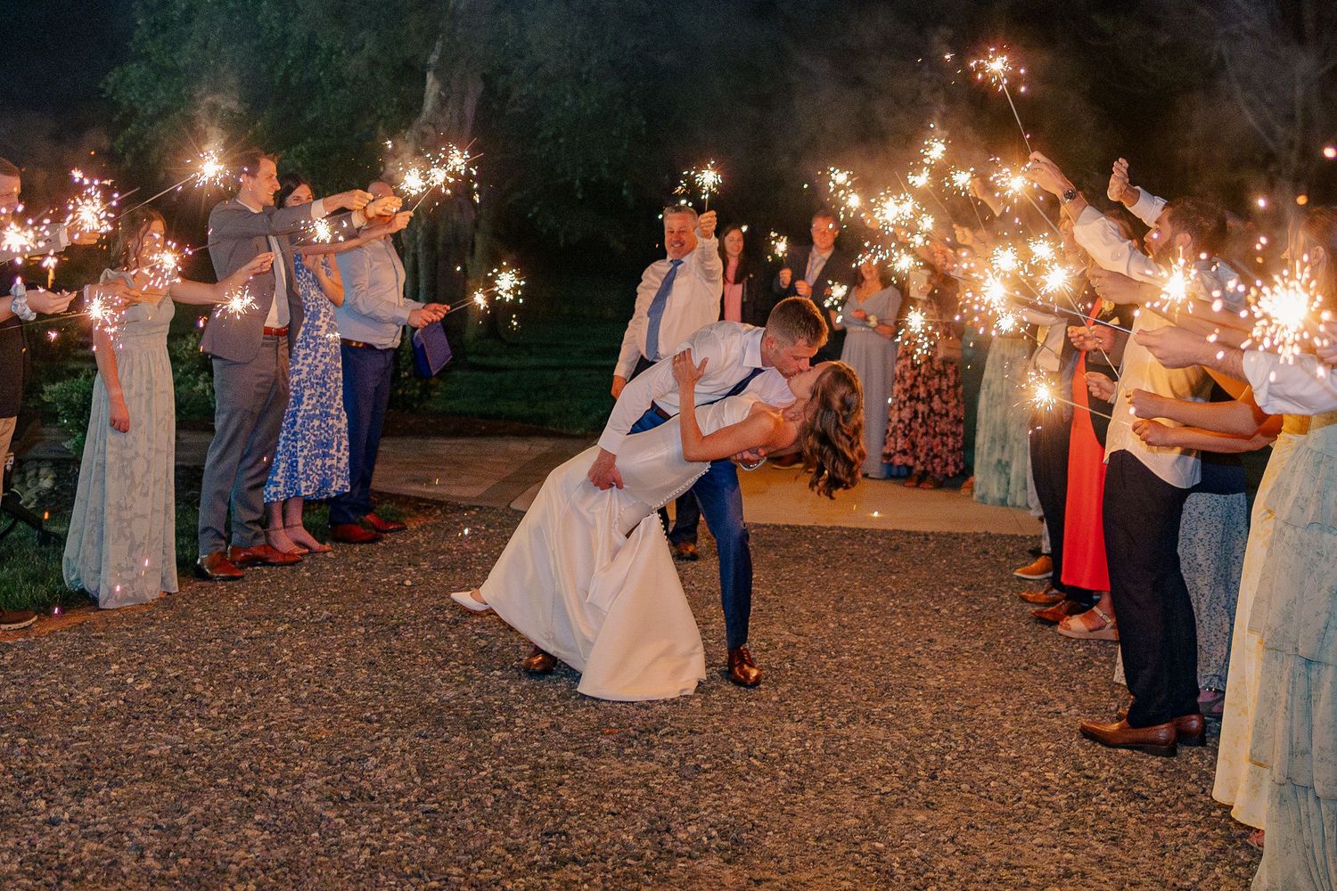 A bride and groom share a romantic dip kiss surrounded by guests holding sparklers at night.