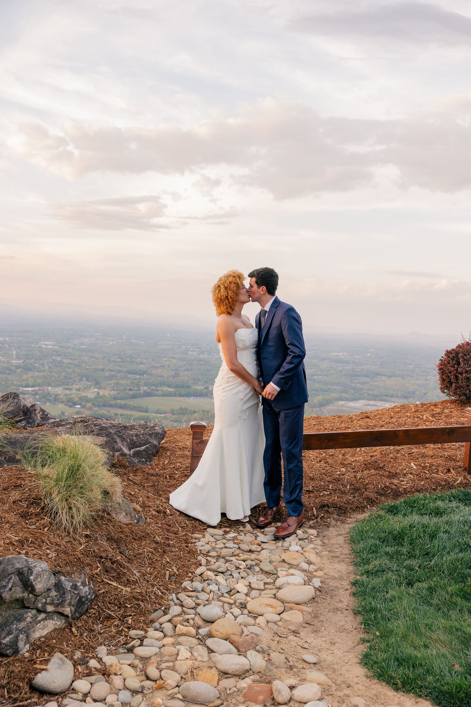 Bride and groom sharing a kiss on a scenic mountain overlook path at sunset.