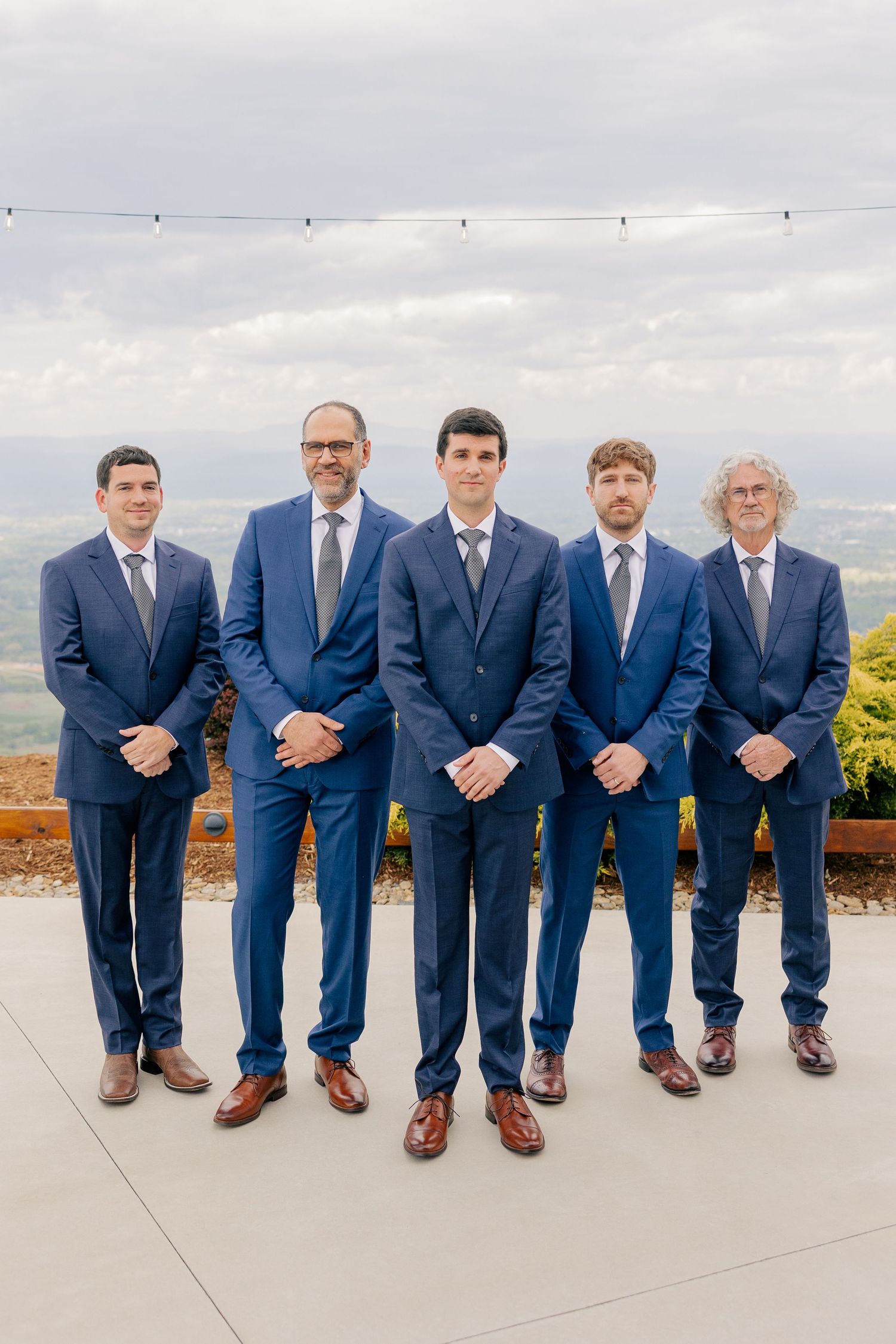 Five groomsmen in matching navy blue suits posing formally on terrace with cloudy sky backdrop.