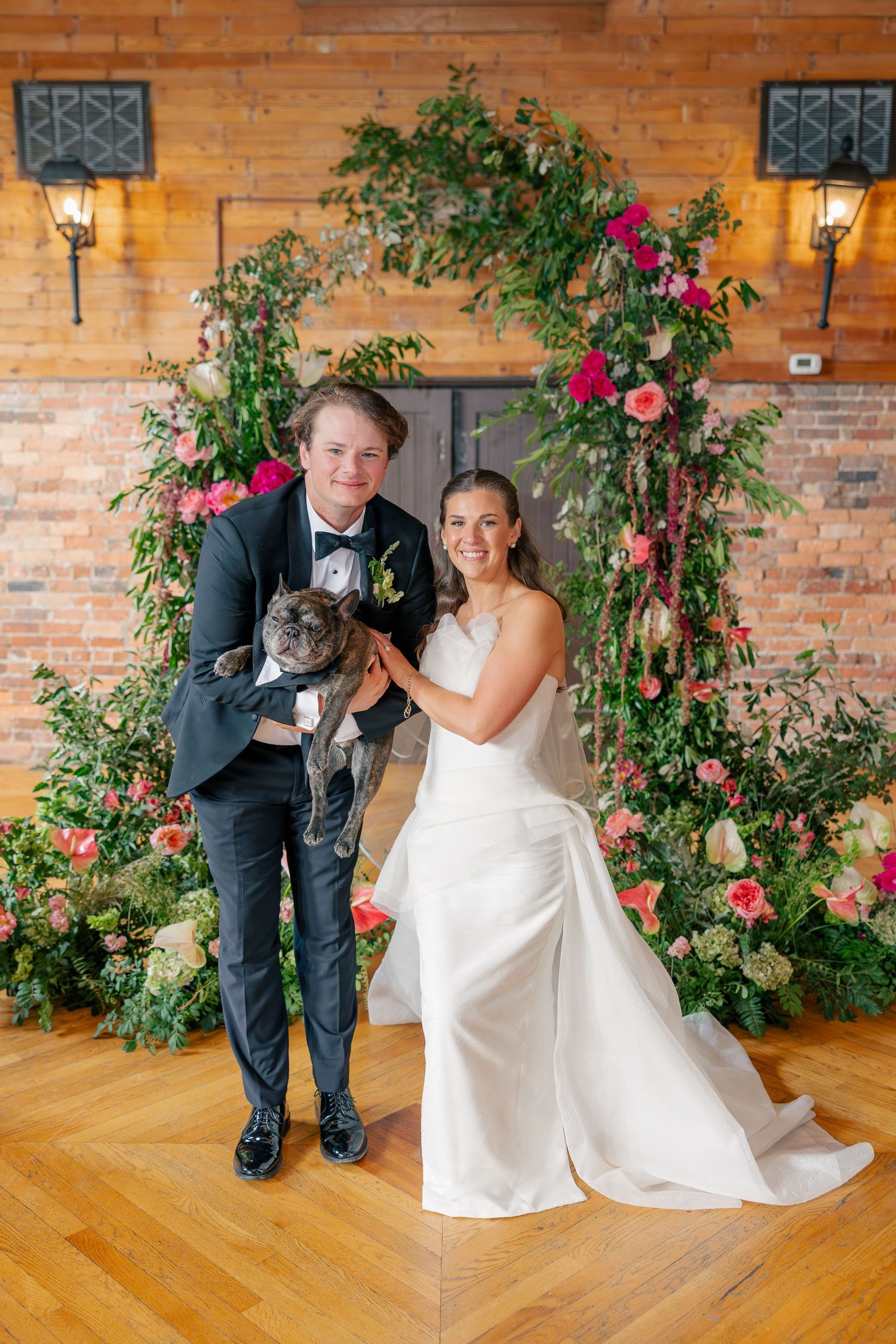 Smiling bride and groom pose with their French bulldog before a lush floral arch in a barn.