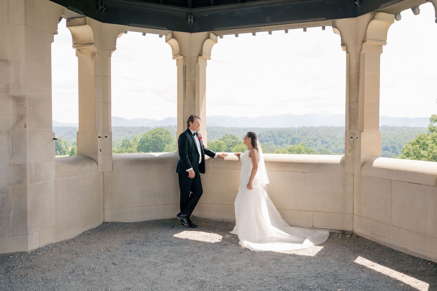 Bride and groom share a romantic moment in an open stone pavilion with scenic landscape views.