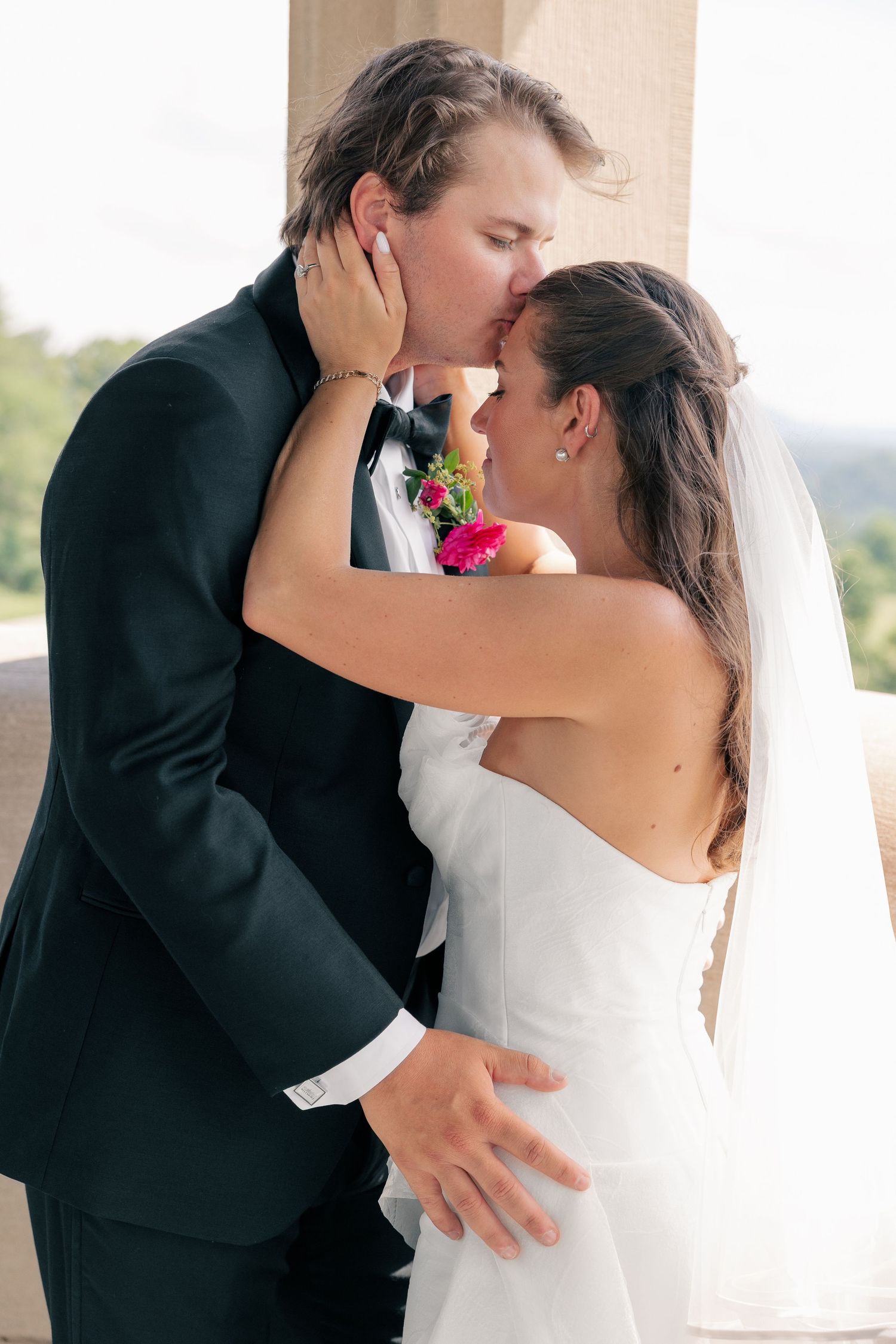 Bride and groom share a tender kiss outdoors between stone columns on their wedding day.