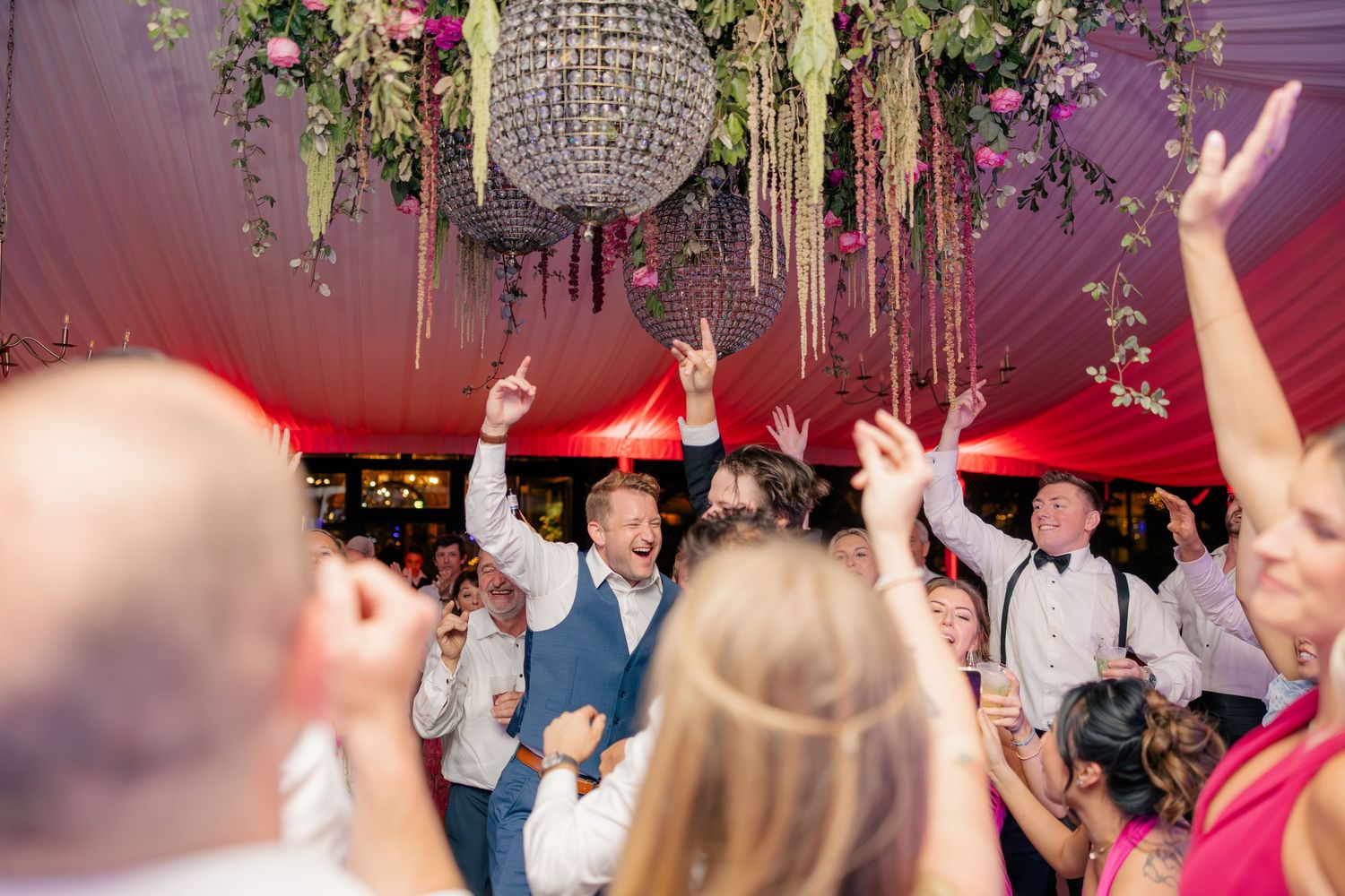 Excited wedding guests celebrate on the dance floor under a crystal chandelier in a decorated tent.