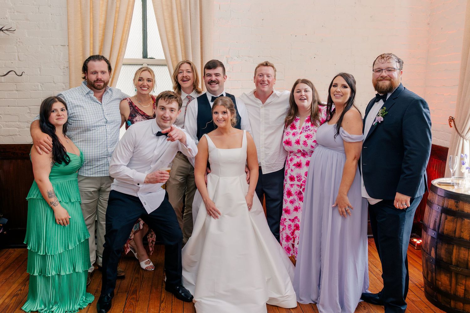 Wedding party group photo with bride in white gown surrounded by guests at indoor reception.
